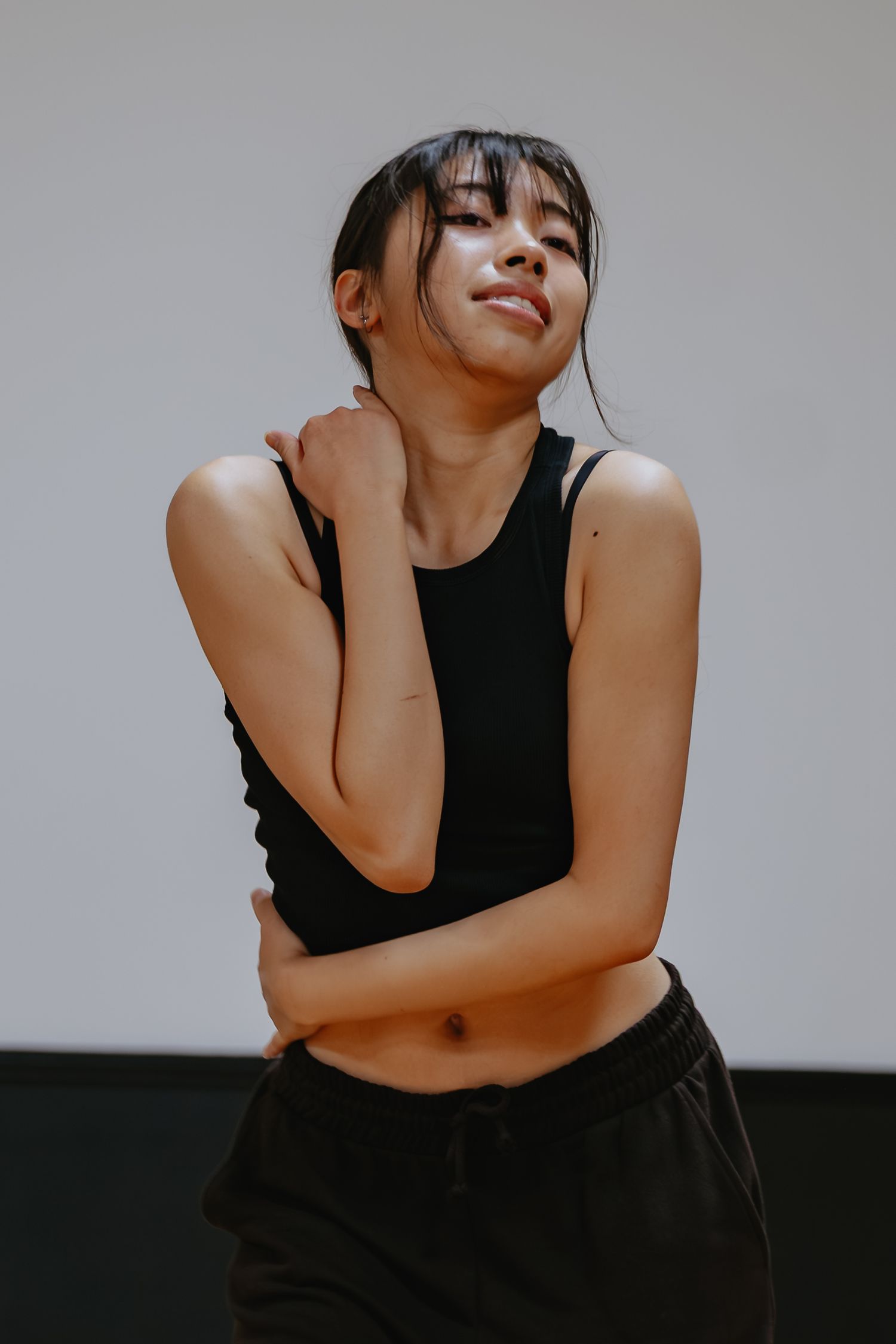 Young woman with dark hair and bangs poses in a black tank top and joggers, arms crossed over her body, smiling in a bright dance studio.