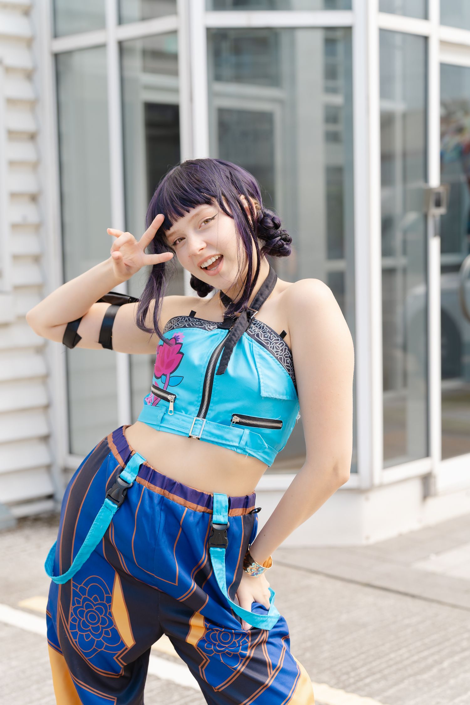 Cosplayer with dark purple hair and bangs poses playfully in a teal crop top and blue patterned pants outside a glass-front building.