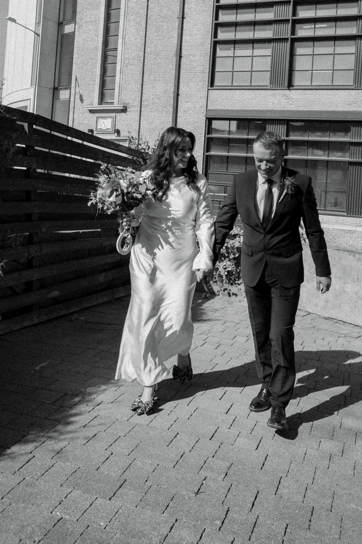 Black and white photo of a smiling bride holding a bouquet walking with her groom on a brick-paved path.