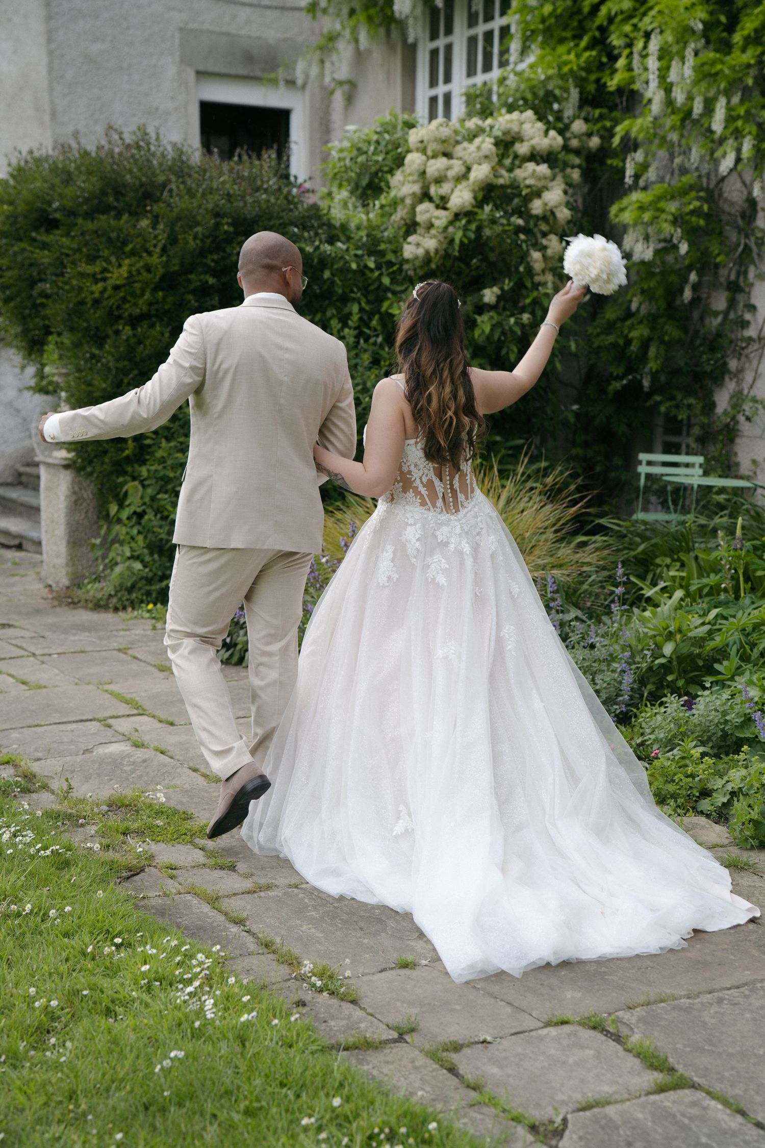 A bride raises her bouquet as she walks with a groom along a stone garden path past flowering shrubs and a white wisteria.