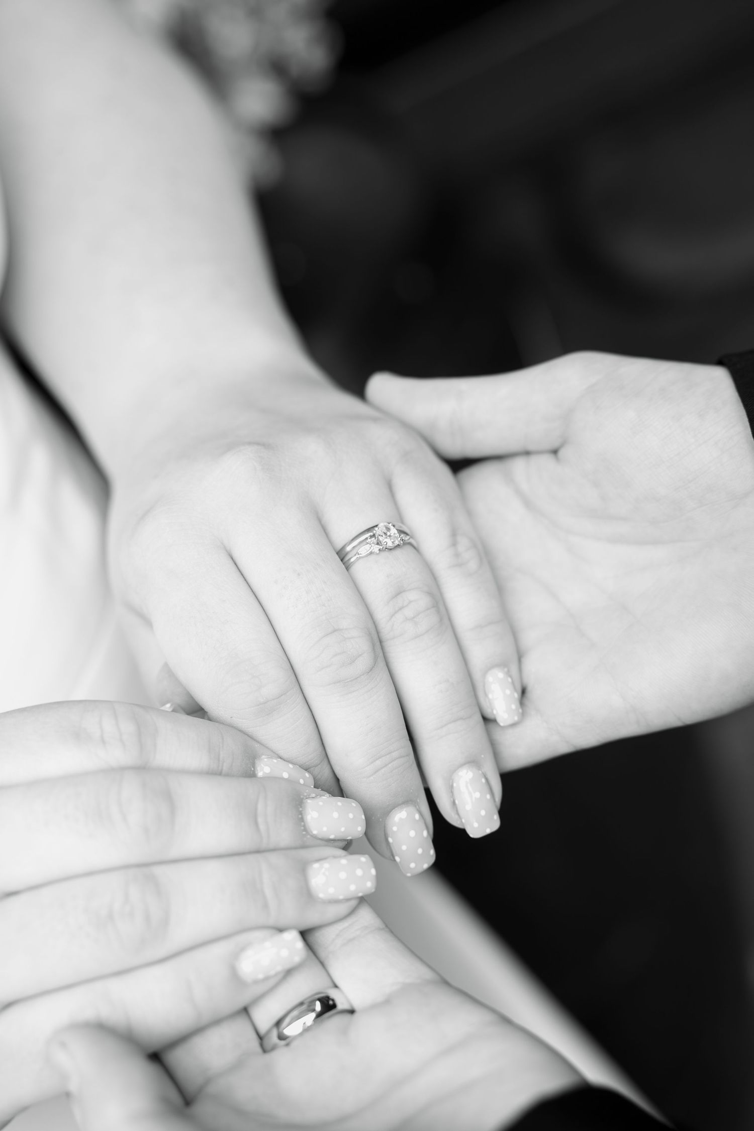 Close-up black and white photo of a couple's joined hands showing a bride's engagement ring, wedding band and polka dot manicure.