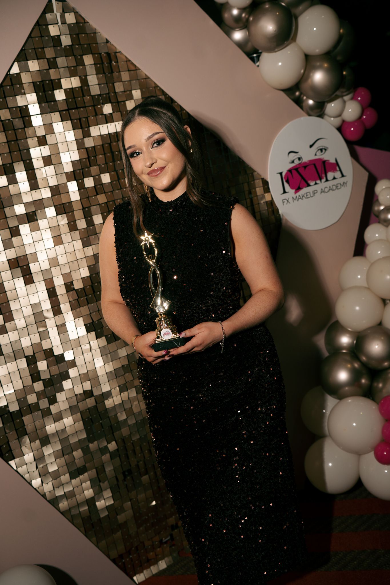 Woman in a sparkly black dress holds a golden award trophy beside a sequin wall and balloon display at a makeup academy event.
