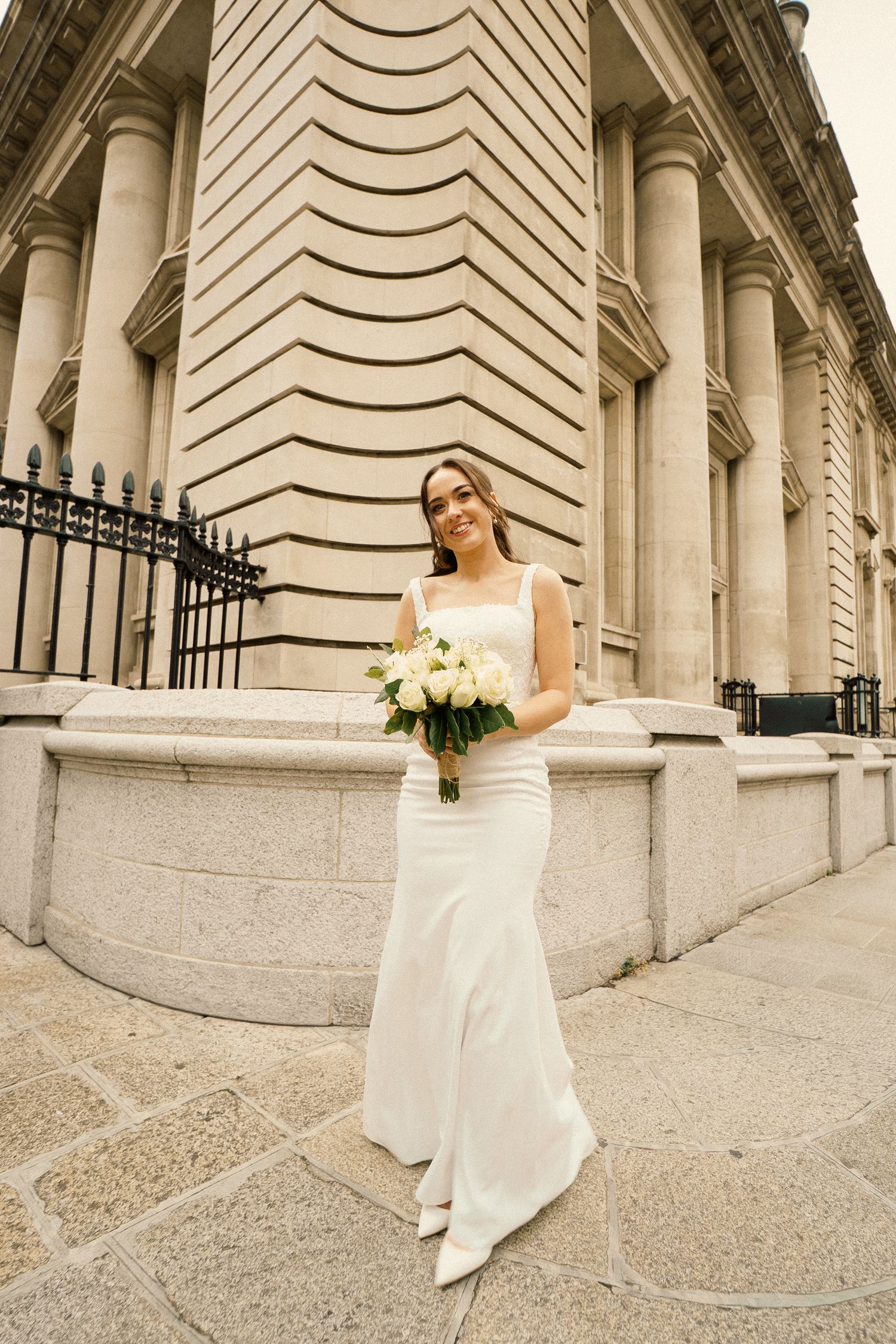 A smiling bride in a sleek white fitted wedding dress holds a white rose bouquet beside a grand stone building with tall columns.
