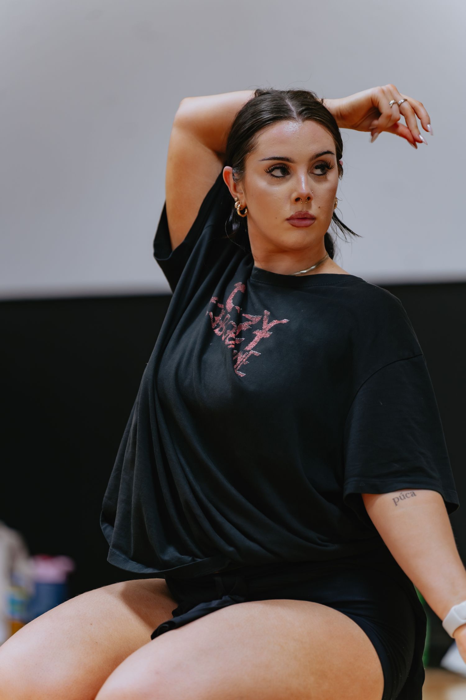 Dancer in a black oversized graphic tee and shorts poses with one arm raised over her head during a studio rehearsal, wearing hoop earrings.