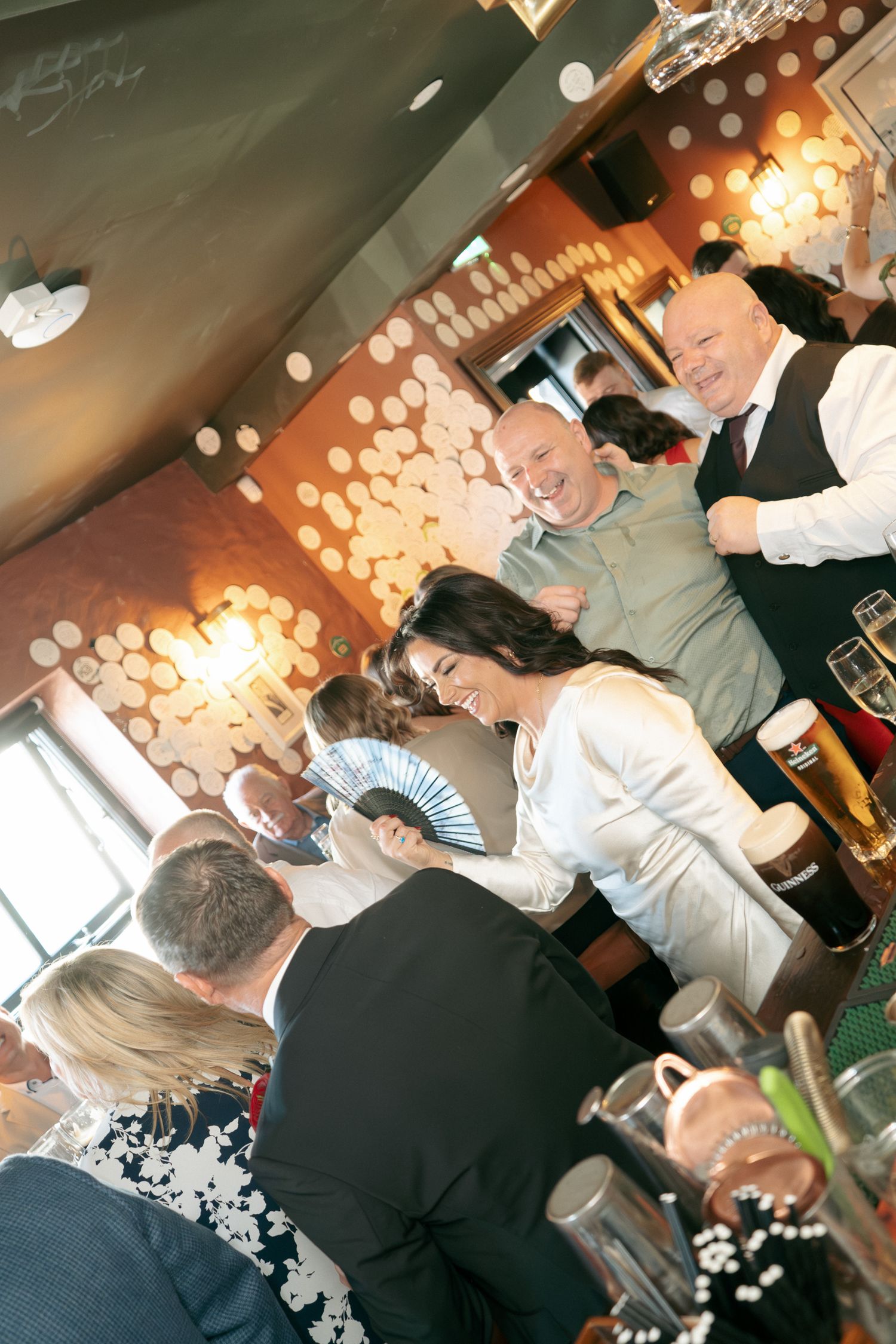 Joyful bride laughs among guests in a lively pub reception with pints of beer, warm lighting and a decorated wall behind them.