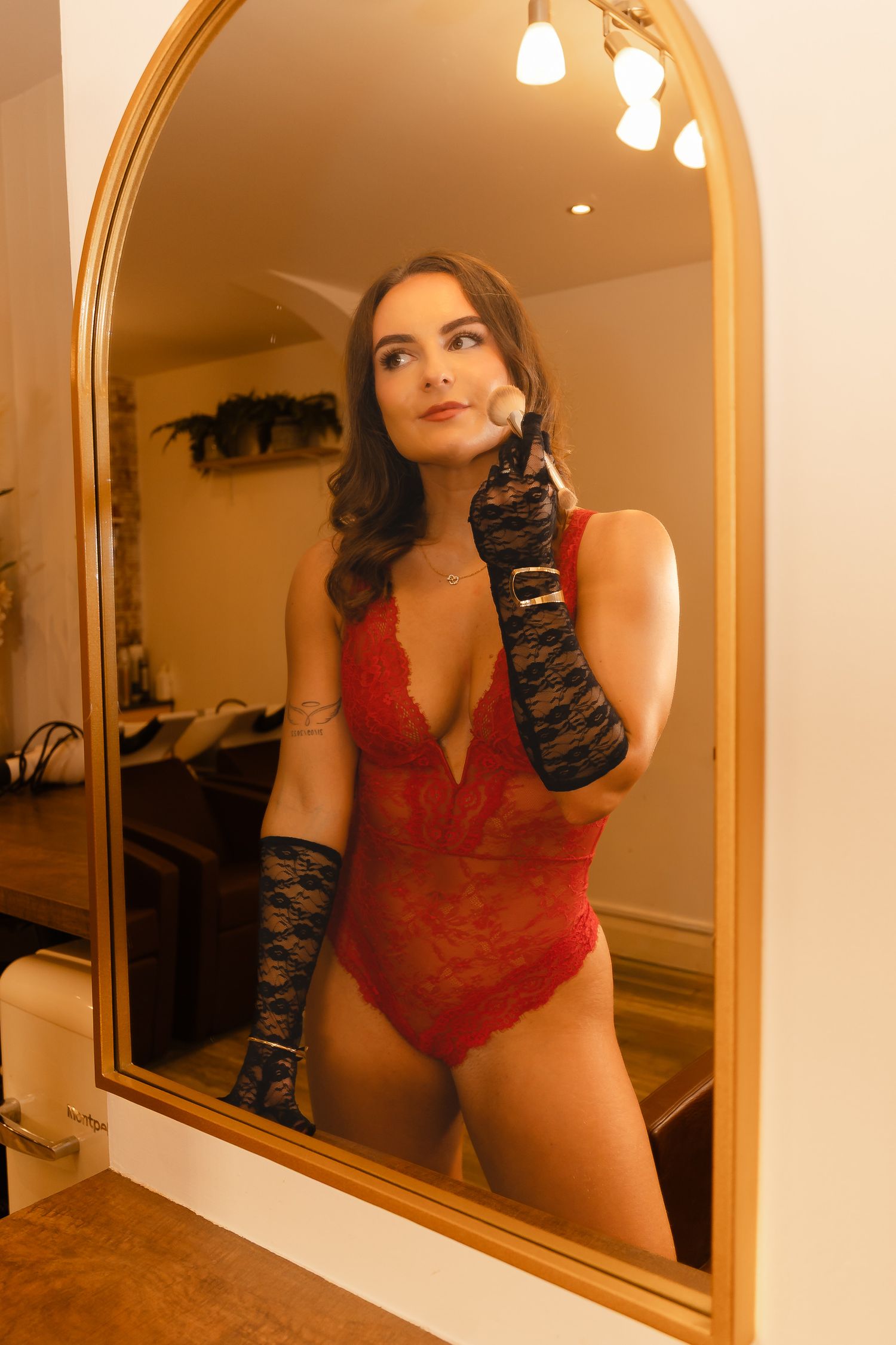 Woman in red lace bodysuit and black lace gloves applies makeup with a brush while looking into a gold arched mirror.