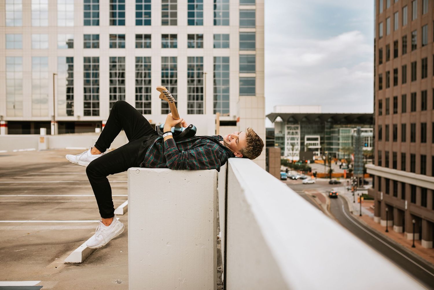 high school senior boy plays guitar on rooftop ledge in orlando cityscape, wearing casual clothes and white sneakers for senior photoshoot.