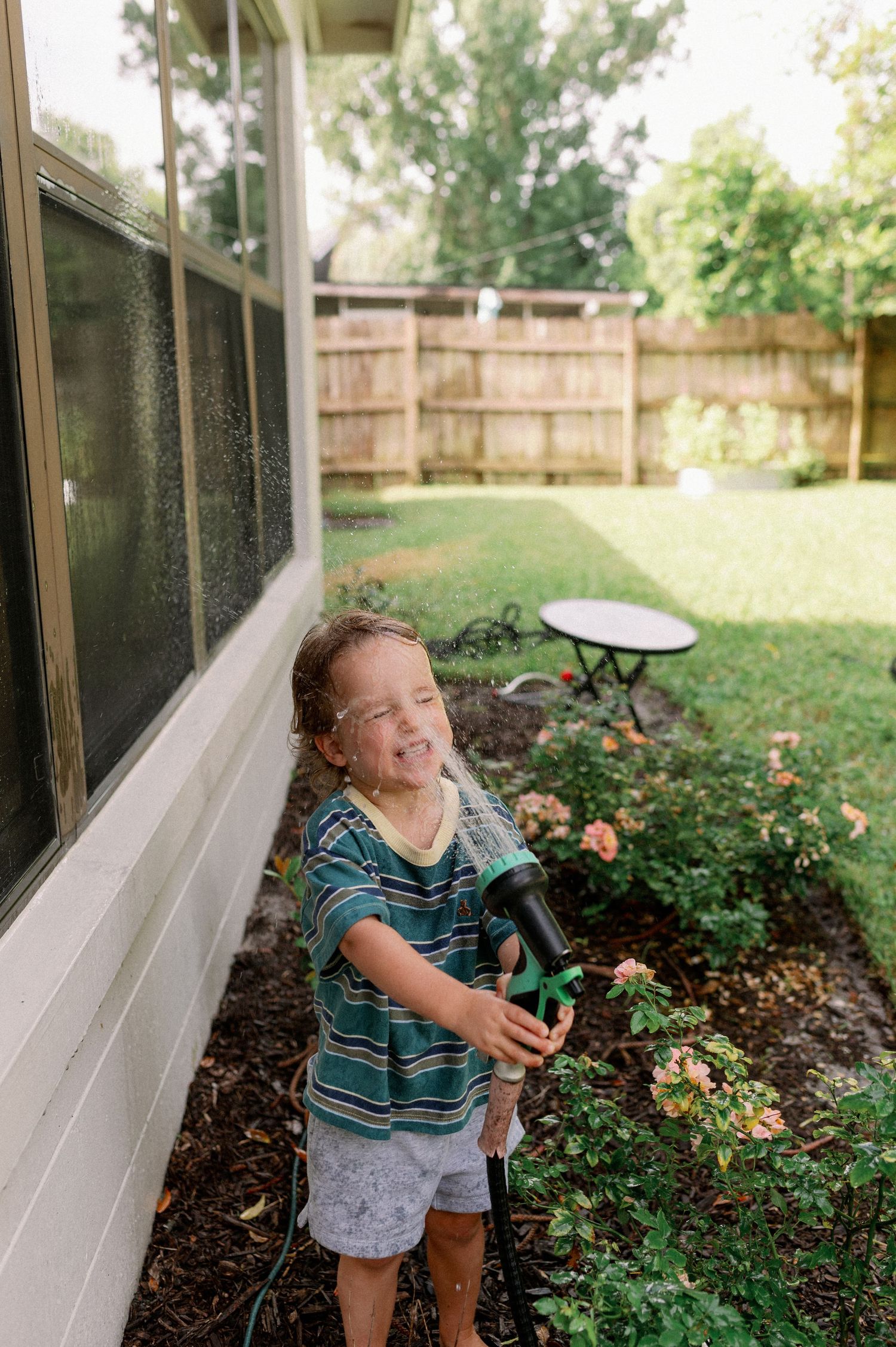 A happy young child holds a garden hose sprayer in a backyard, getting wet while watering near a house window during an in-home photo shoot