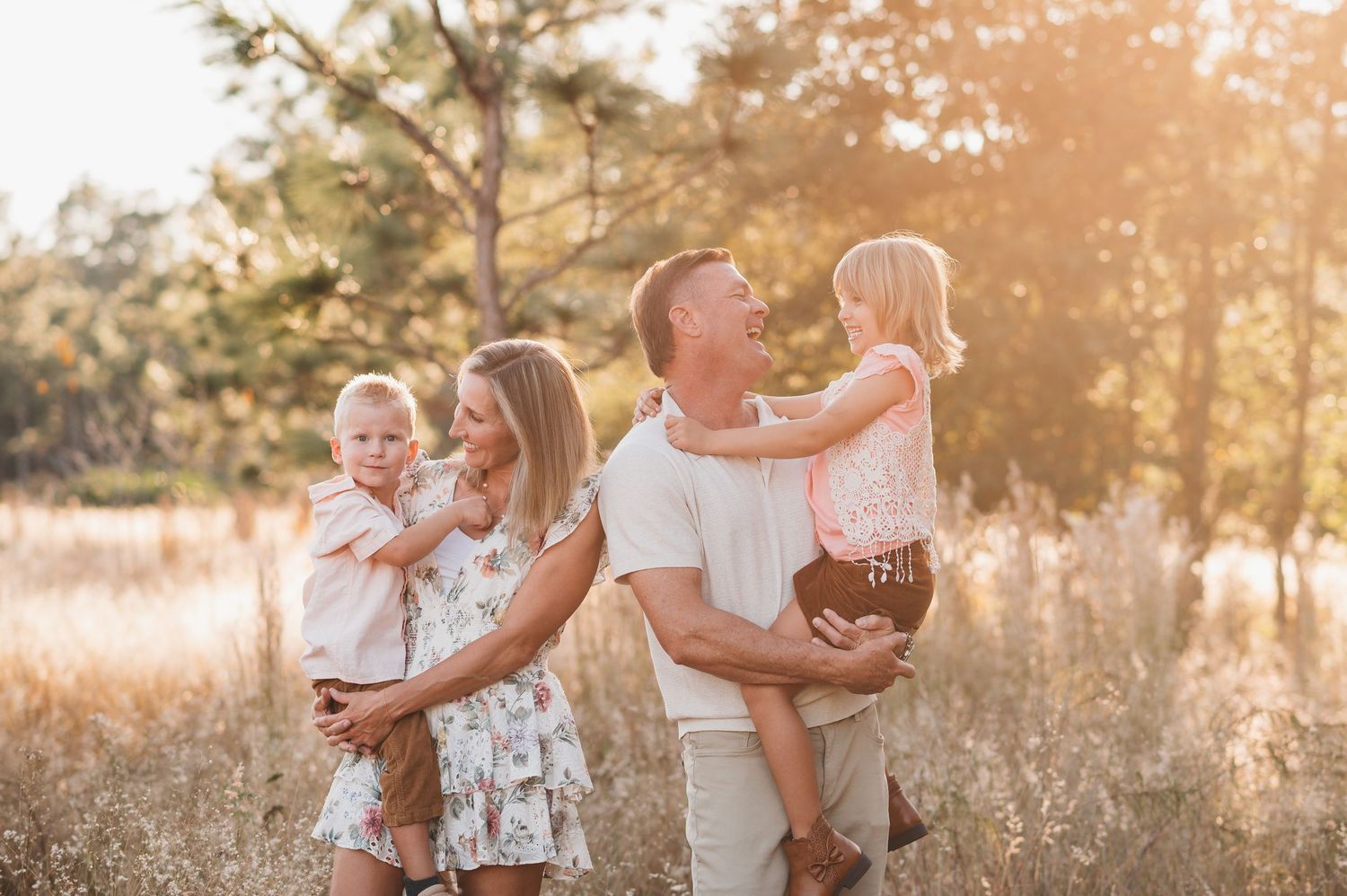 Happy family of four enjoying a golden hour outdoor photo session in a sunlit meadow with tall grass and trees in Clermont fl
