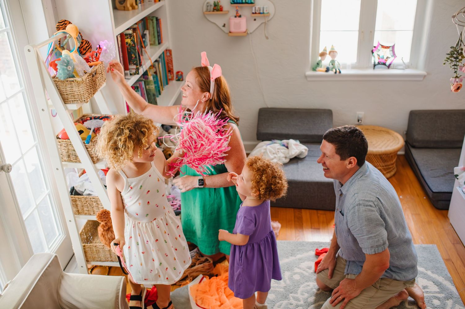 Family with young children playing and decorating in a bright living room with bookshelves and hardwood floors during an in-home photo shoot