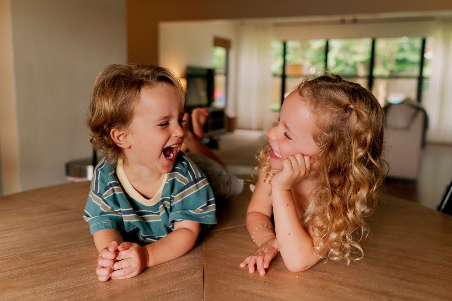 Two young children laughing and smiling together on a wooden table in a cozy home interior during an in-home photo session in orlando fl