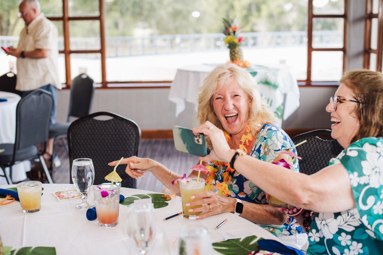Event photographer captures two women in tropical attire laughing at a decorated table with colorful cocktails at a festive party.