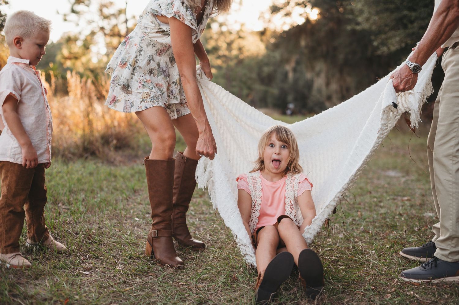 Family photo session outdoors with two young children, parents swinging daughter in white lace blanket, golden hour lighting at Lake Louisa