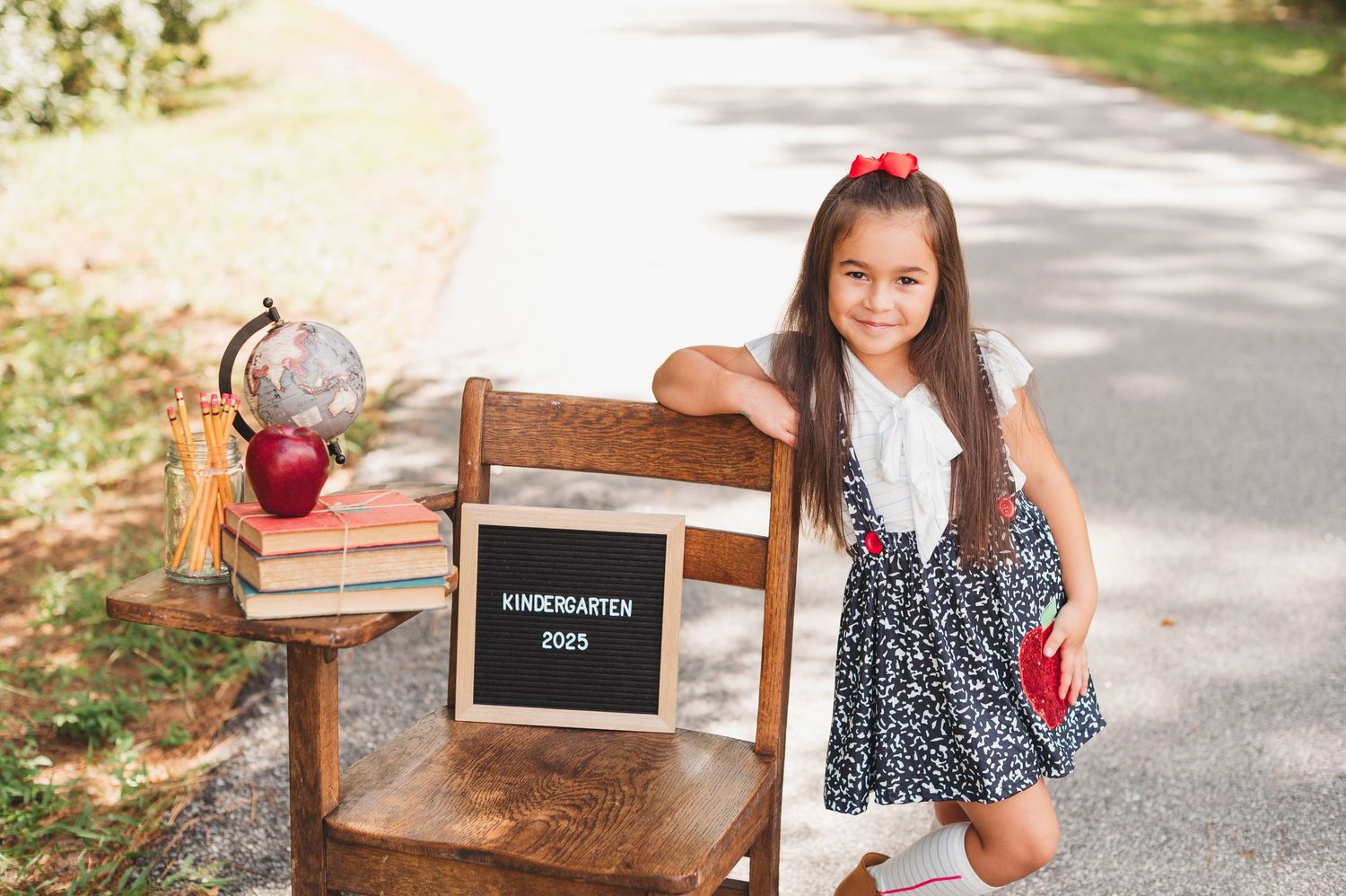 Girl poses beside a wooden chair with a Kindergarten 2025 sign, books, globe, and apple for a back-to-school photo in sanford fl
