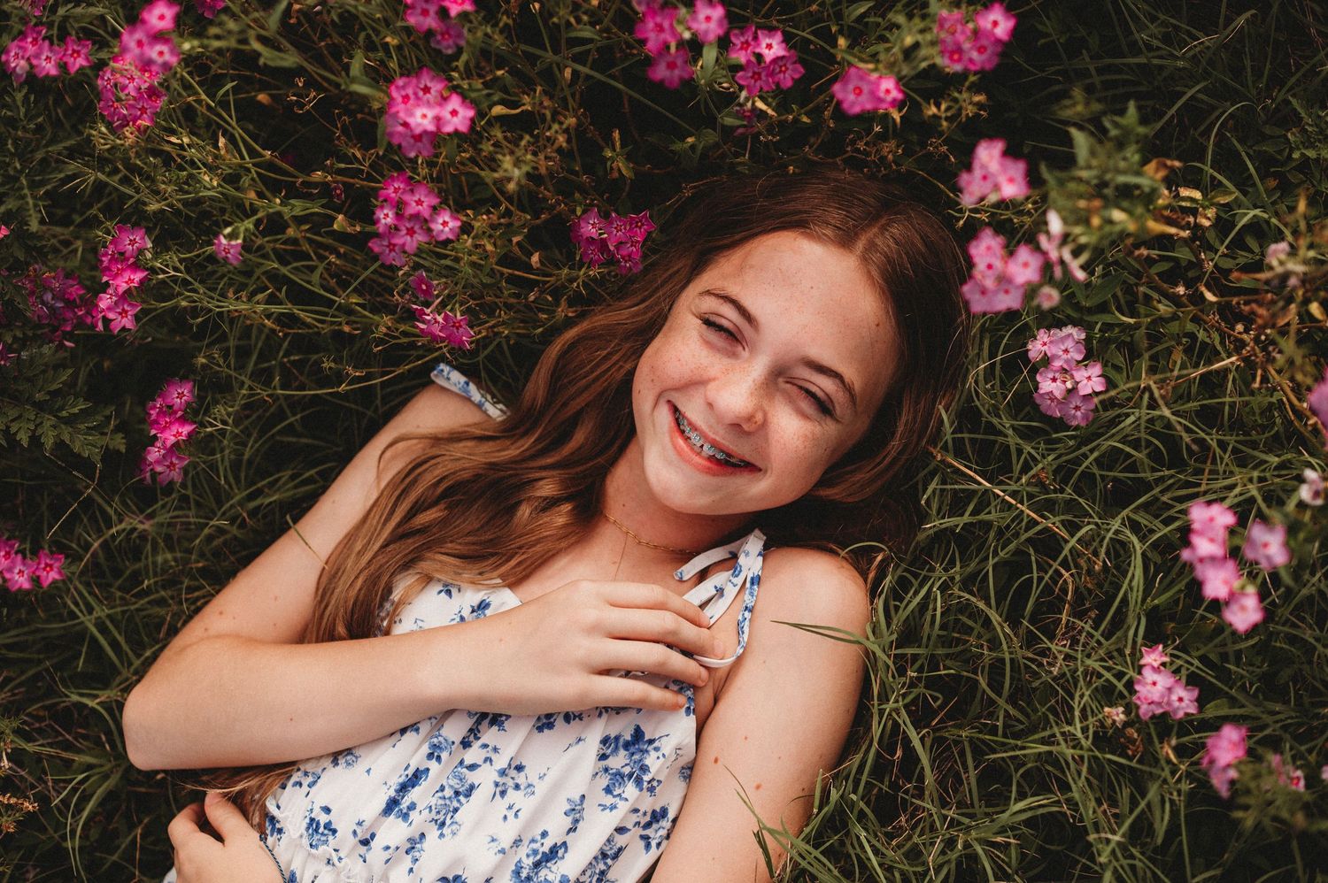 A smiling teenage girl lies in green grass surrounded by pink flowers wearing a floral dress during a mommy and me shoot in apopka florida