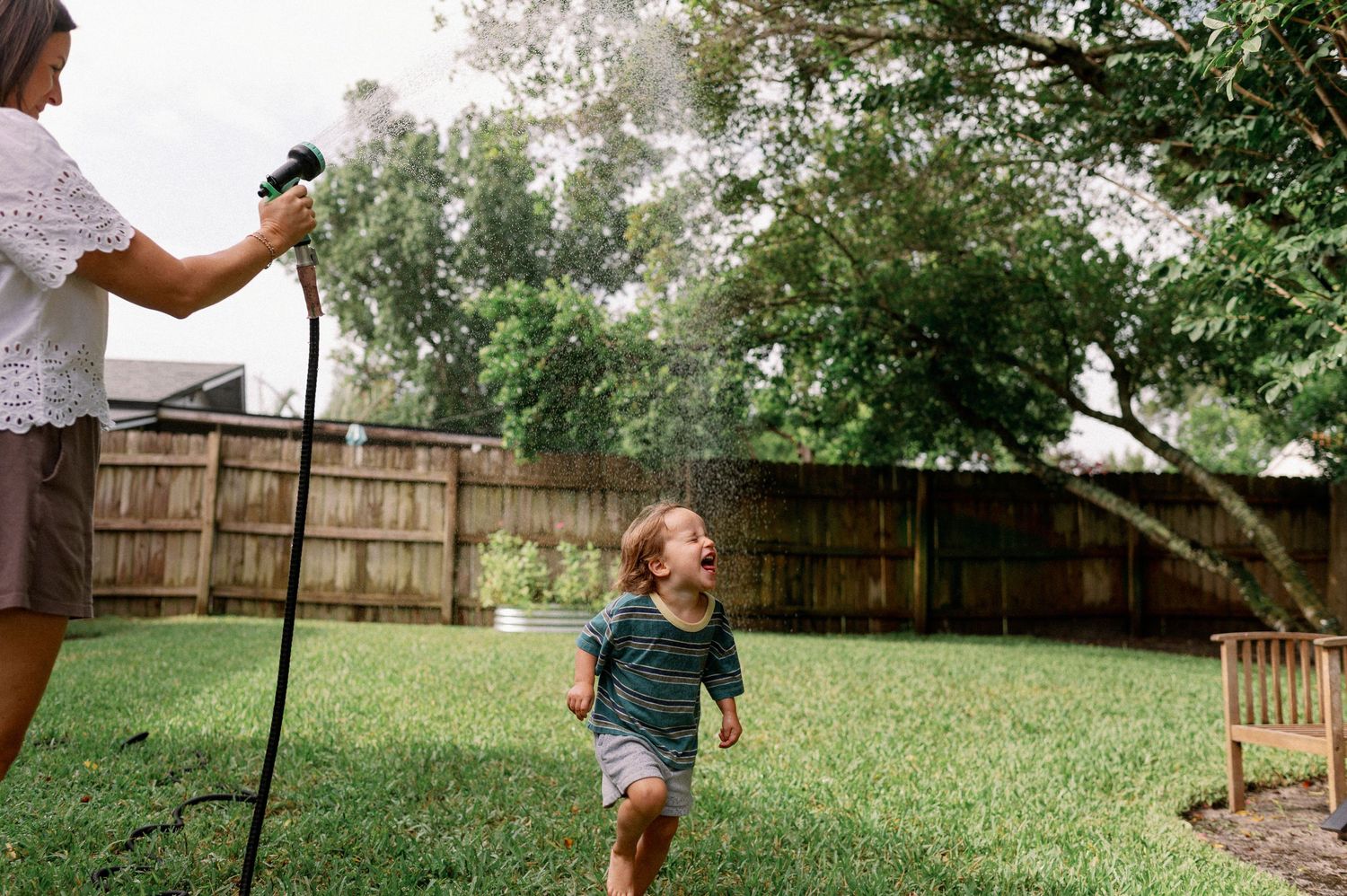 Child laughing and running through backyard sprinkler while adult sprays water hose on sunny day during an in-home photo session in orlando