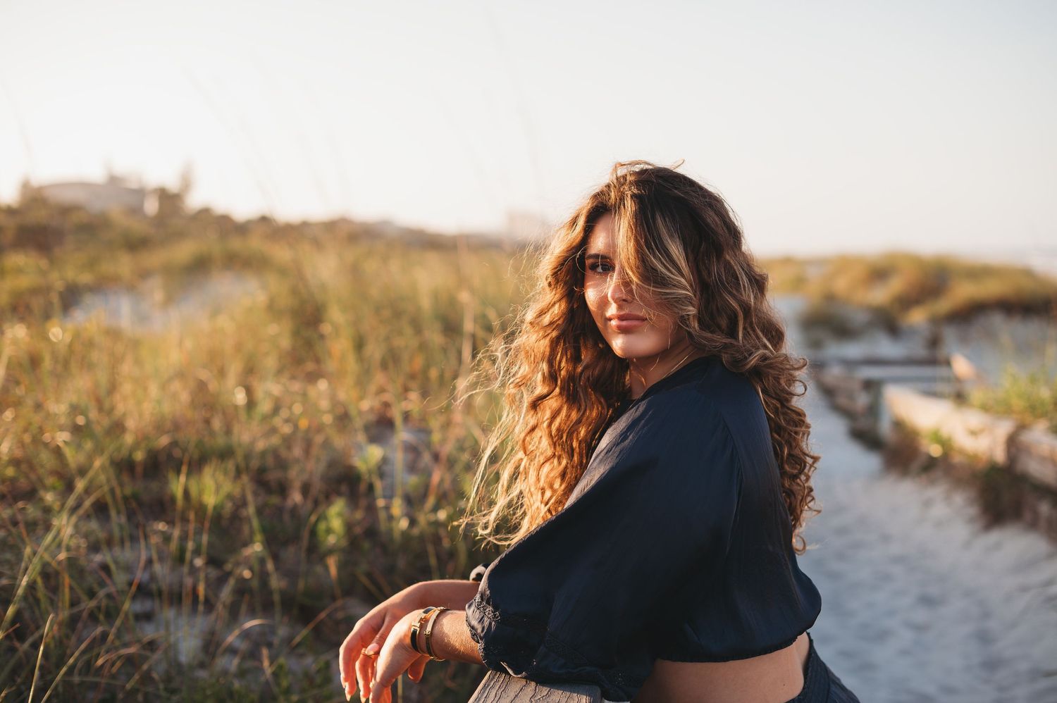 high school senior girl poses on NSB beach path at golden hour, wearing a dark top with tall grass behind her for senior photos.