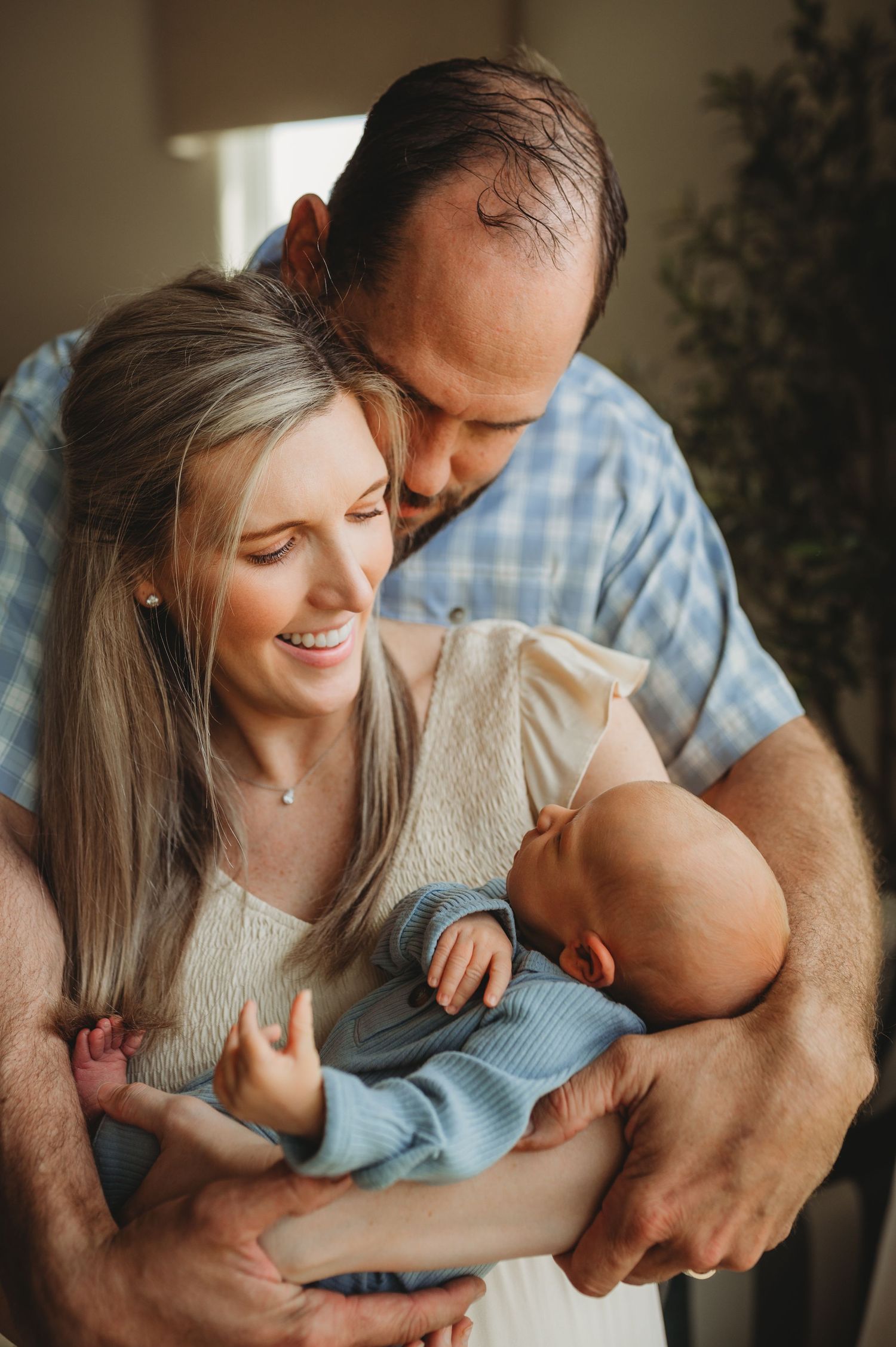 A smiling couple tenderly holds their newborn baby, with the father kissing the mother's head in an in-home newborn session in orlando