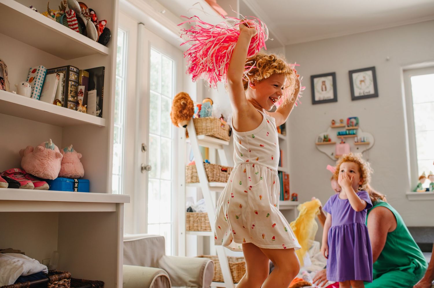 Two young girls playing in a bright, toy-filled bedroom, one dancing with a pink feather duster during an orlando in-home photo session.