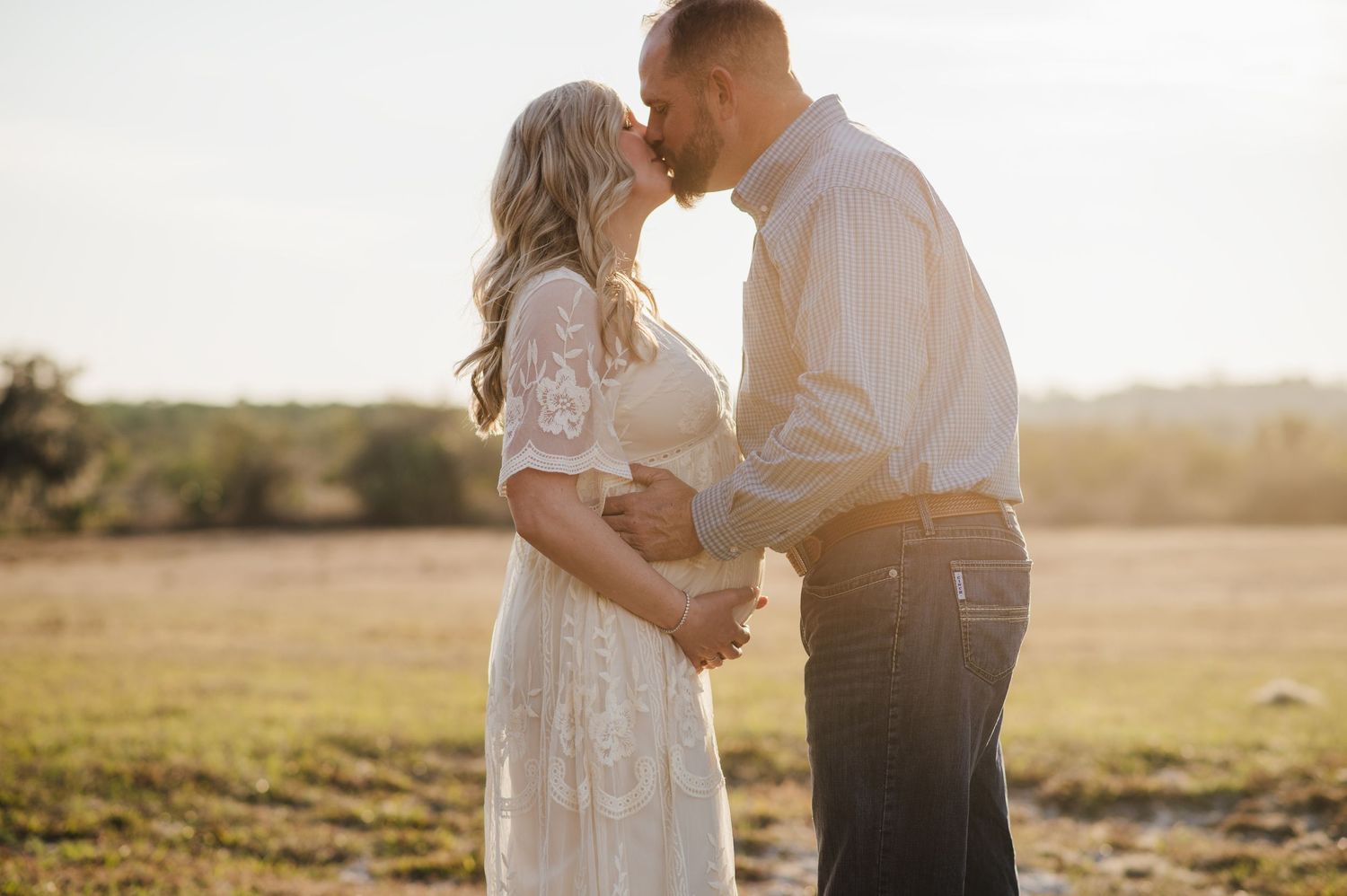 Couple sharing a kiss outdoors at golden hour, woman in white lace maternity dress cradling baby bump in open field for a fl maternity shoot
