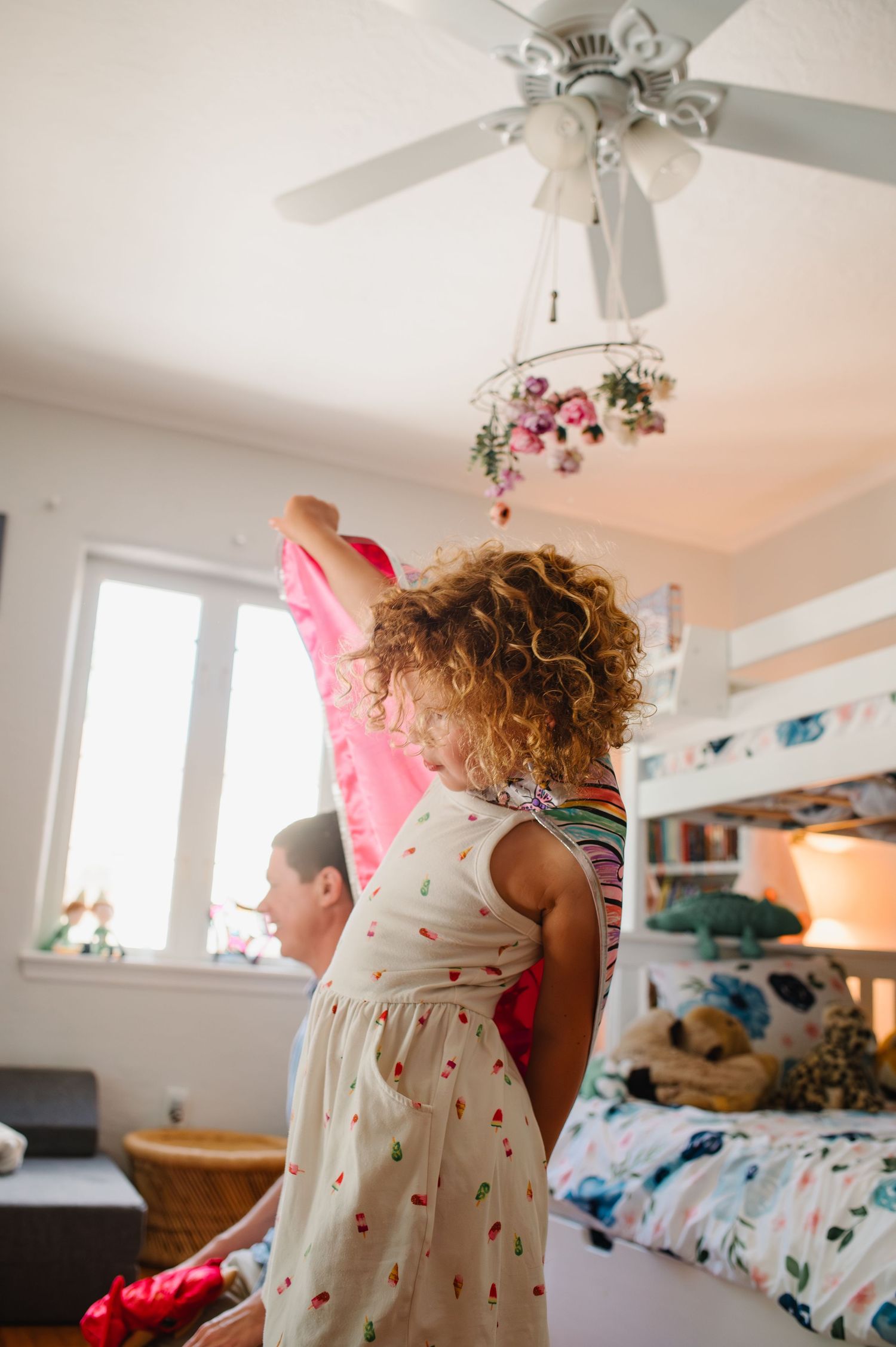 Young girl with curly hair in floral dress reaches up toward ceiling fan in bright kids bedroom with bunk bed during an in-home photo shoot.
