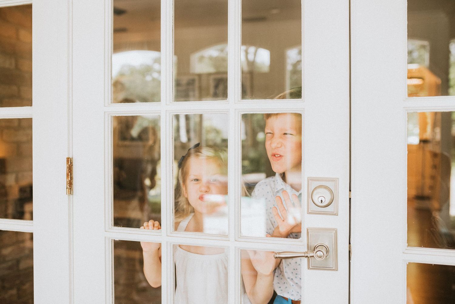 Two children peering through white French doors with glass panes in a bright home entryway during an in-home photography session in orlando
