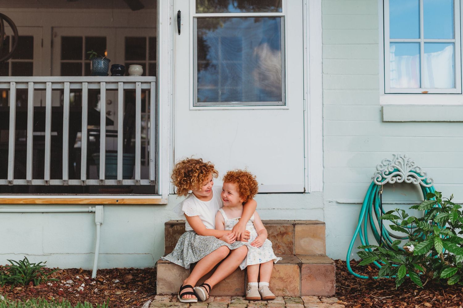 Two curly-haired sisters sit together on front porch steps of a light green house, sharing a sweet moment during an in-home family shoot.