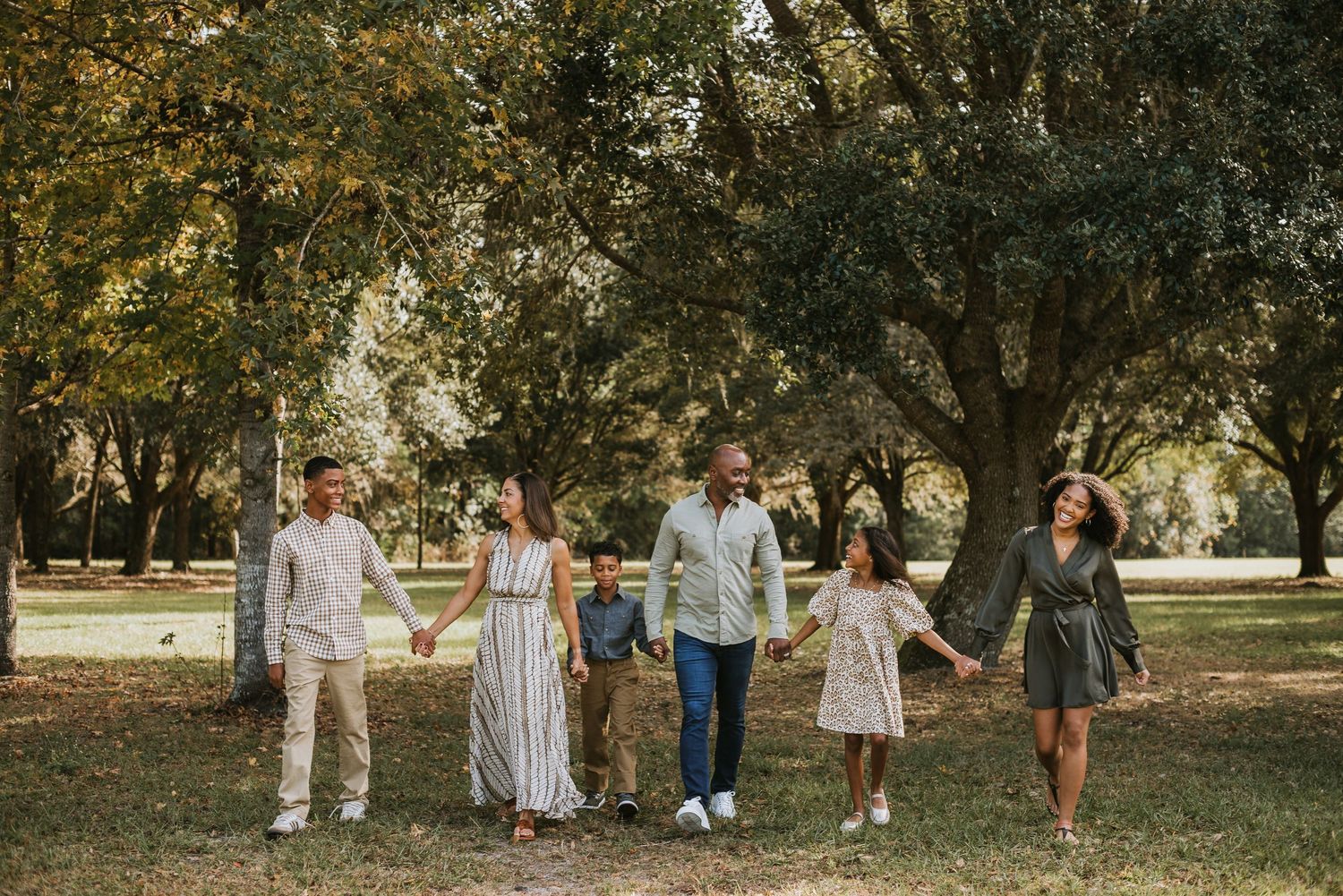 Family of six holding hands walking through a lush green park under large oak trees on a sunny day for family photos in sanford fl