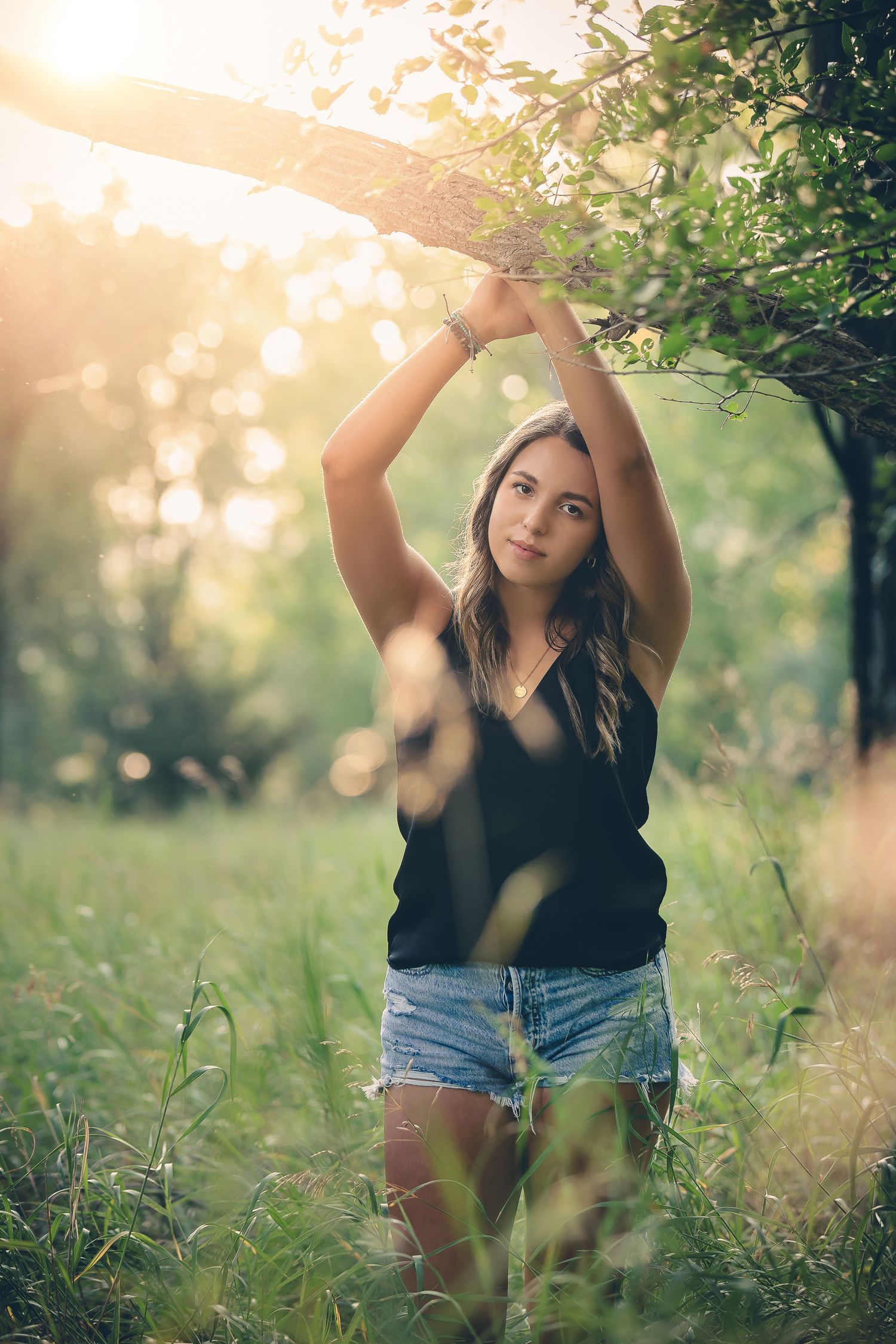 Summer portrait in black tank top and denim shorts reaching up to tree branches.