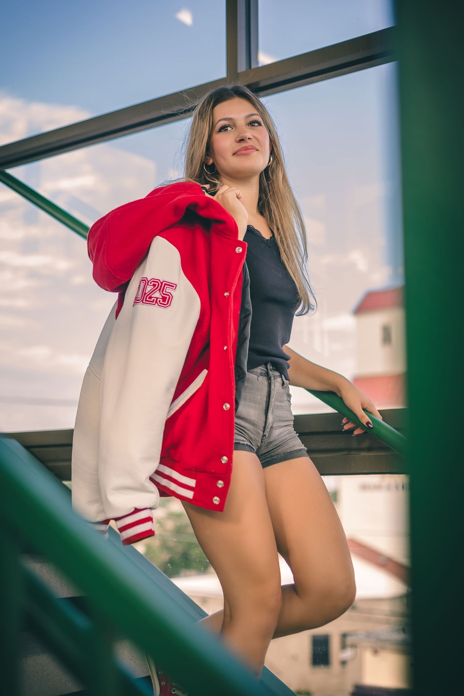 Fashion portrait on stairs wearing varsity jacket and shorts against blue sky.