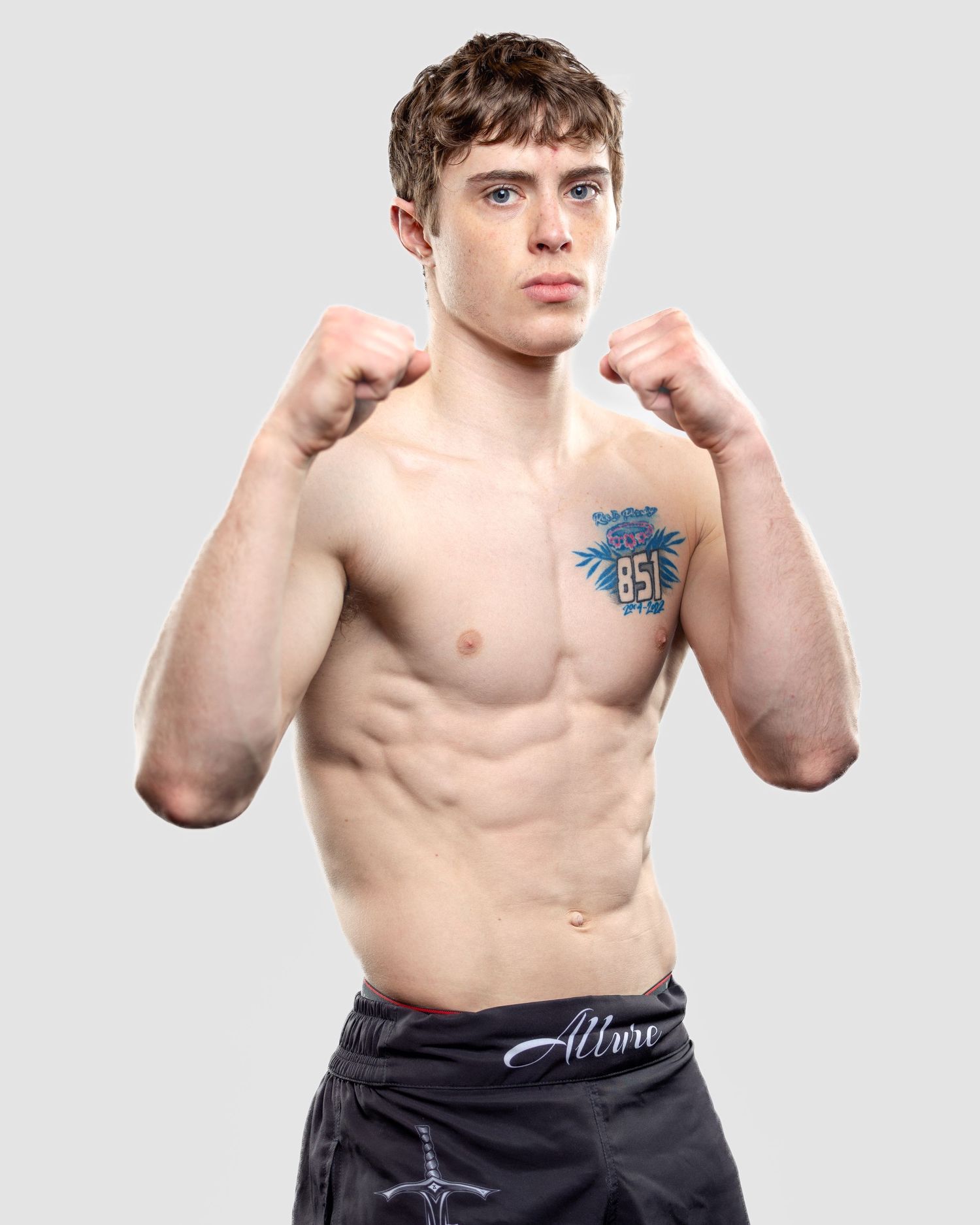 Shirtless athlete in black shorts poses in boxing stance showing torso and blue tattoo on chest against grey background.