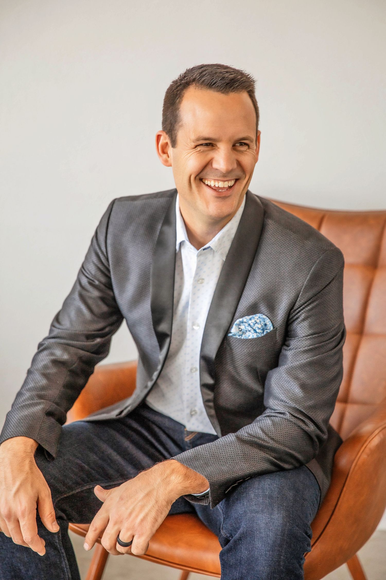 A professional in a gray blazer with blue pocket square smiles while sitting in a brown leather chair.