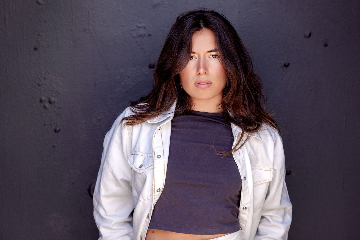 Person wearing a white denim jacket and navy crop top poses confidently against dark textured wall.