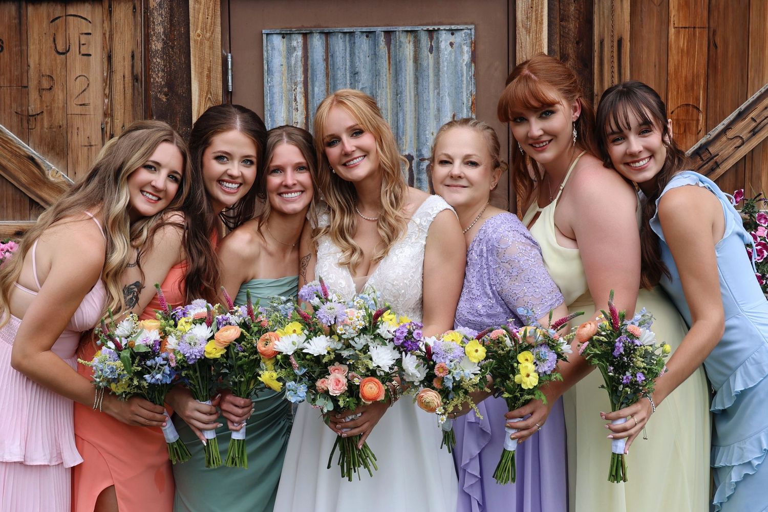 Bride with five bridesmaids in colorful dresses holding wildflower bouquets posing in front of rustic barn doors.