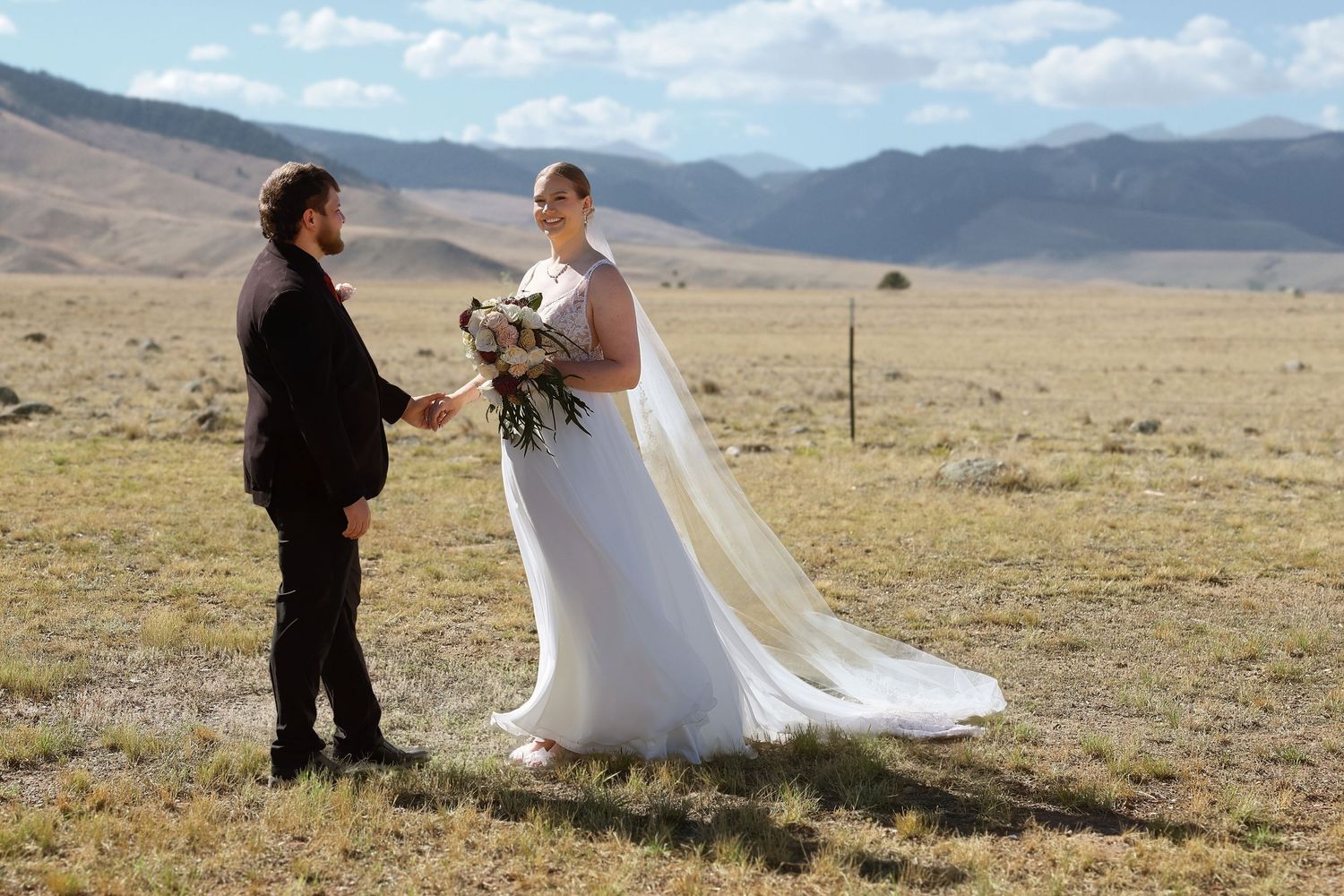 Bride and groom holding hands in a vast open field with mountain backdrop on their wedding day.