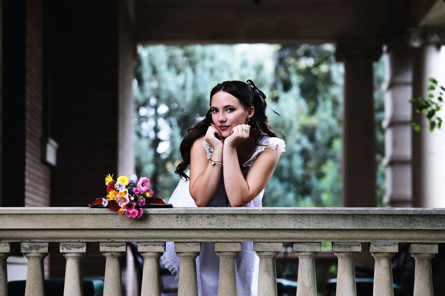 Bride in white dress leaning on stone balcony railing with colorful bouquet nearby.