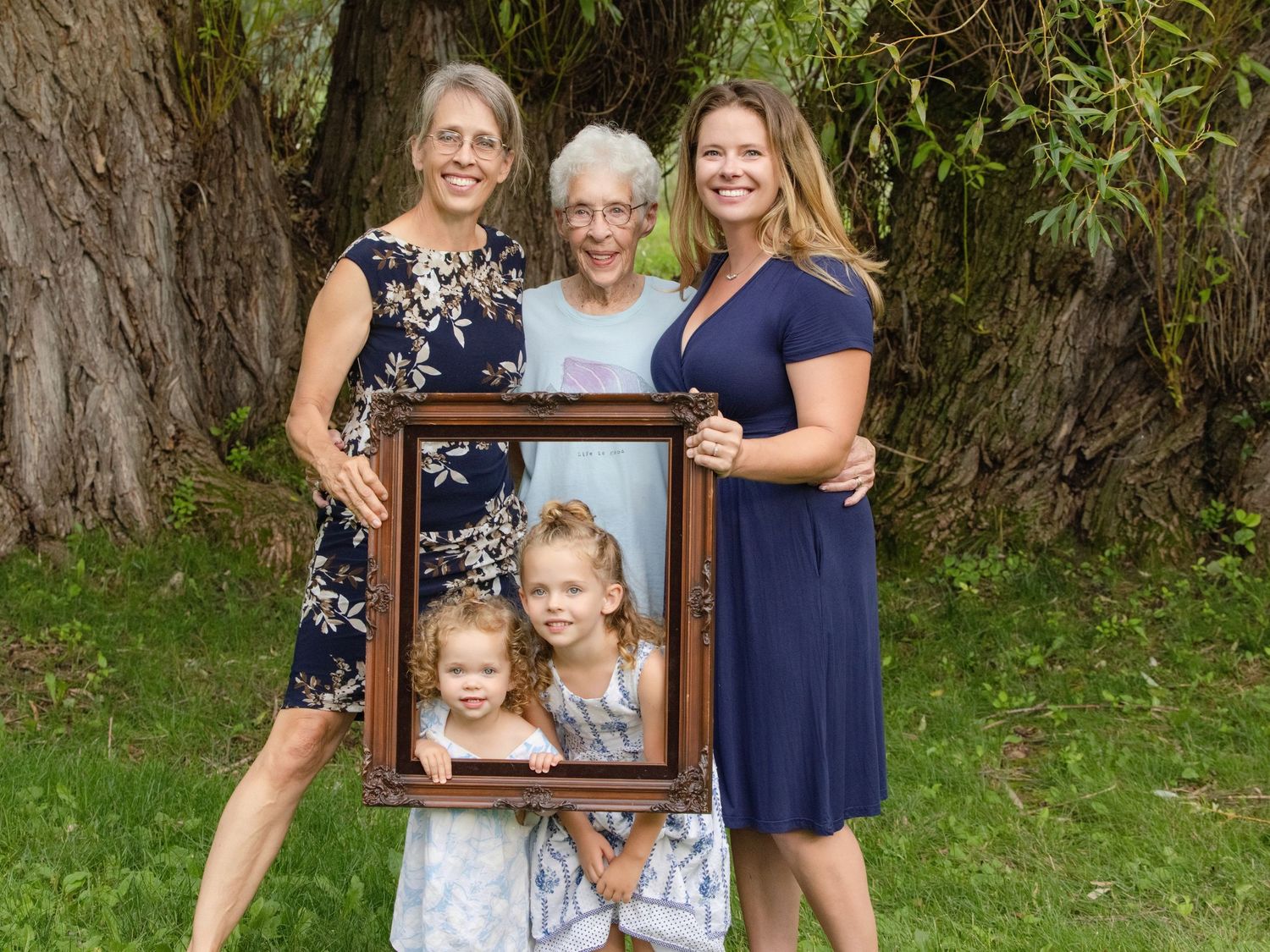 Three generations pose outdoors holding a framed photo of two young children in front of a large tree.
