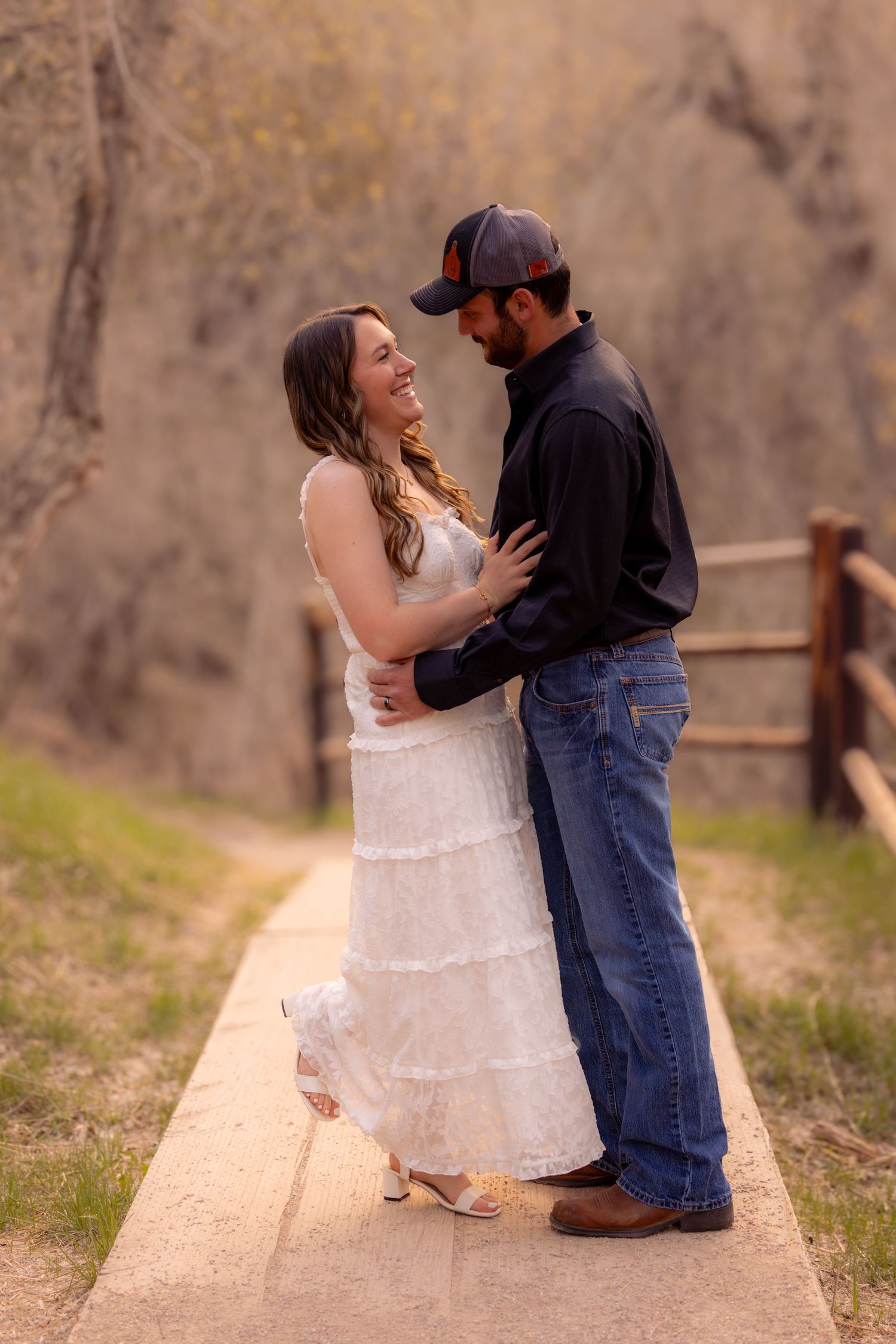 Couple embracing on a path, woman in white dress and man in jeans and cap, smiling in a natural outdoor setting.