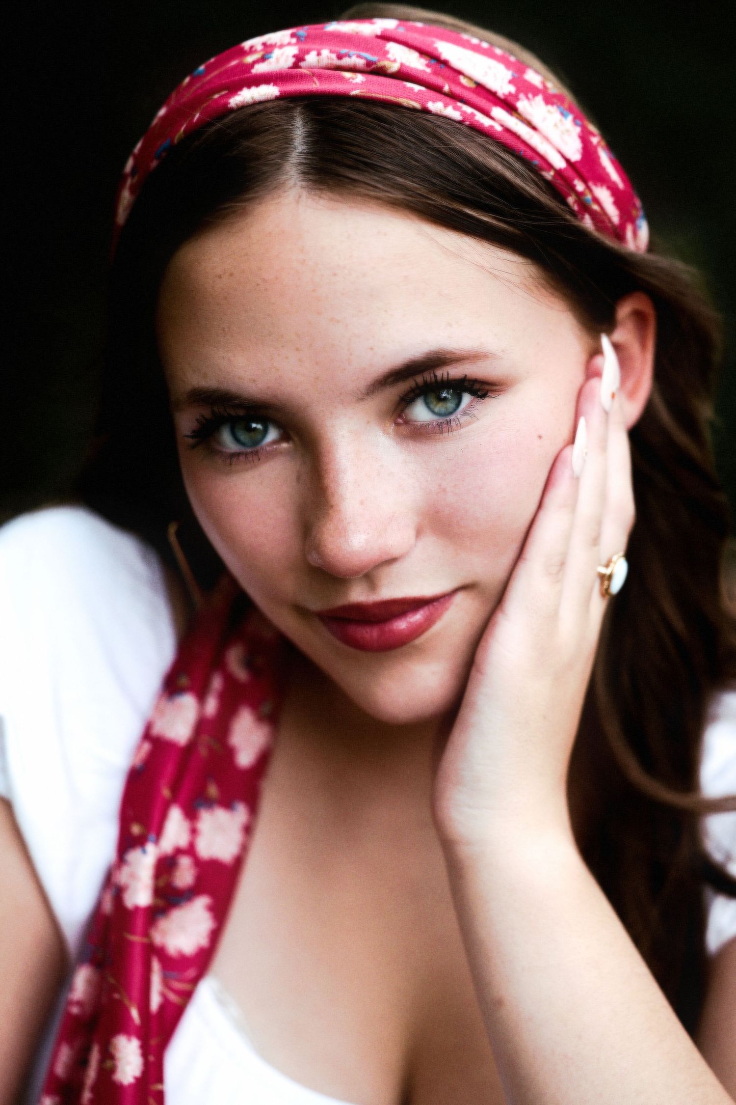 Young woman with green eyes, red floral headband, and dark lip color resting hand on cheek.