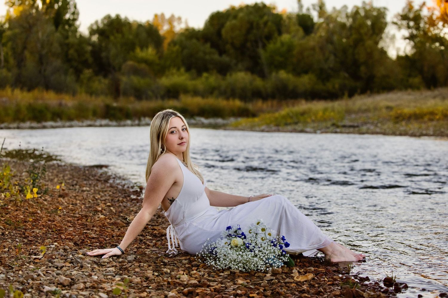 Blonde woman in white dress sitting by a river with wildflowers, surrounded by lush green trees at golden hour.