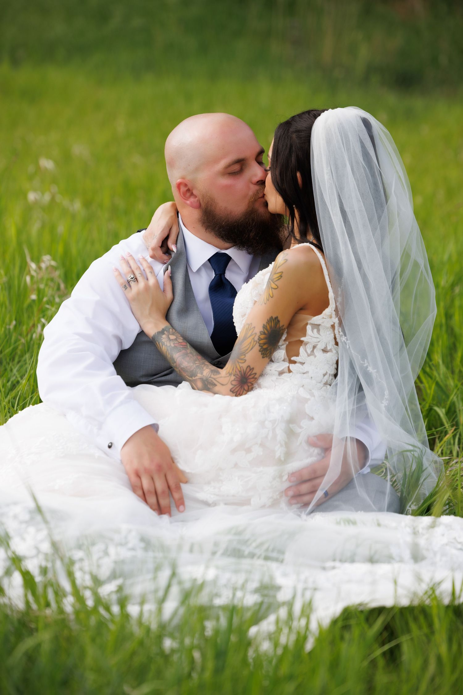 Bride and groom sharing a kiss while sitting in a lush green field, bride wearing a white lace gown and veil.