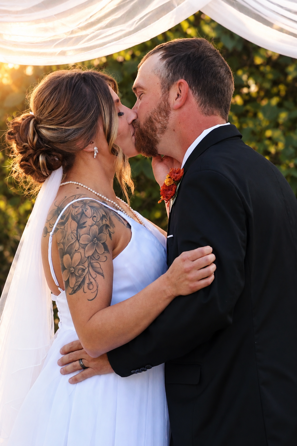A bride and groom share their first kiss at an outdoor wedding ceremony, with golden sunset light glowing behind them.