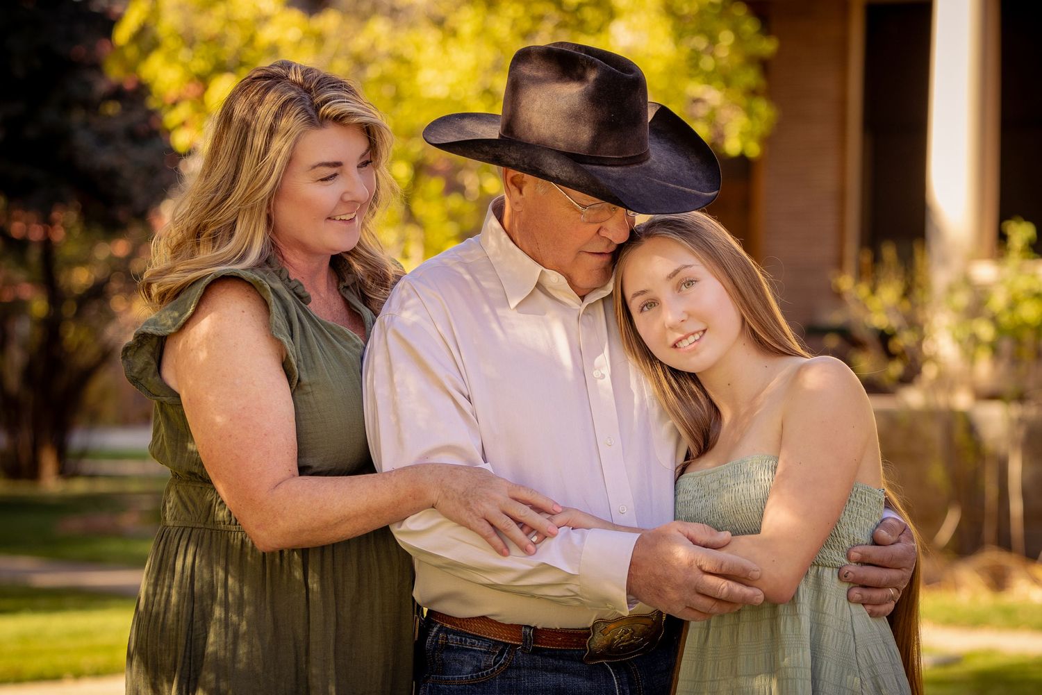 A family of three shares a warm embrace outdoors, with a man in a cowboy hat hugging two women in a sunny garden.
