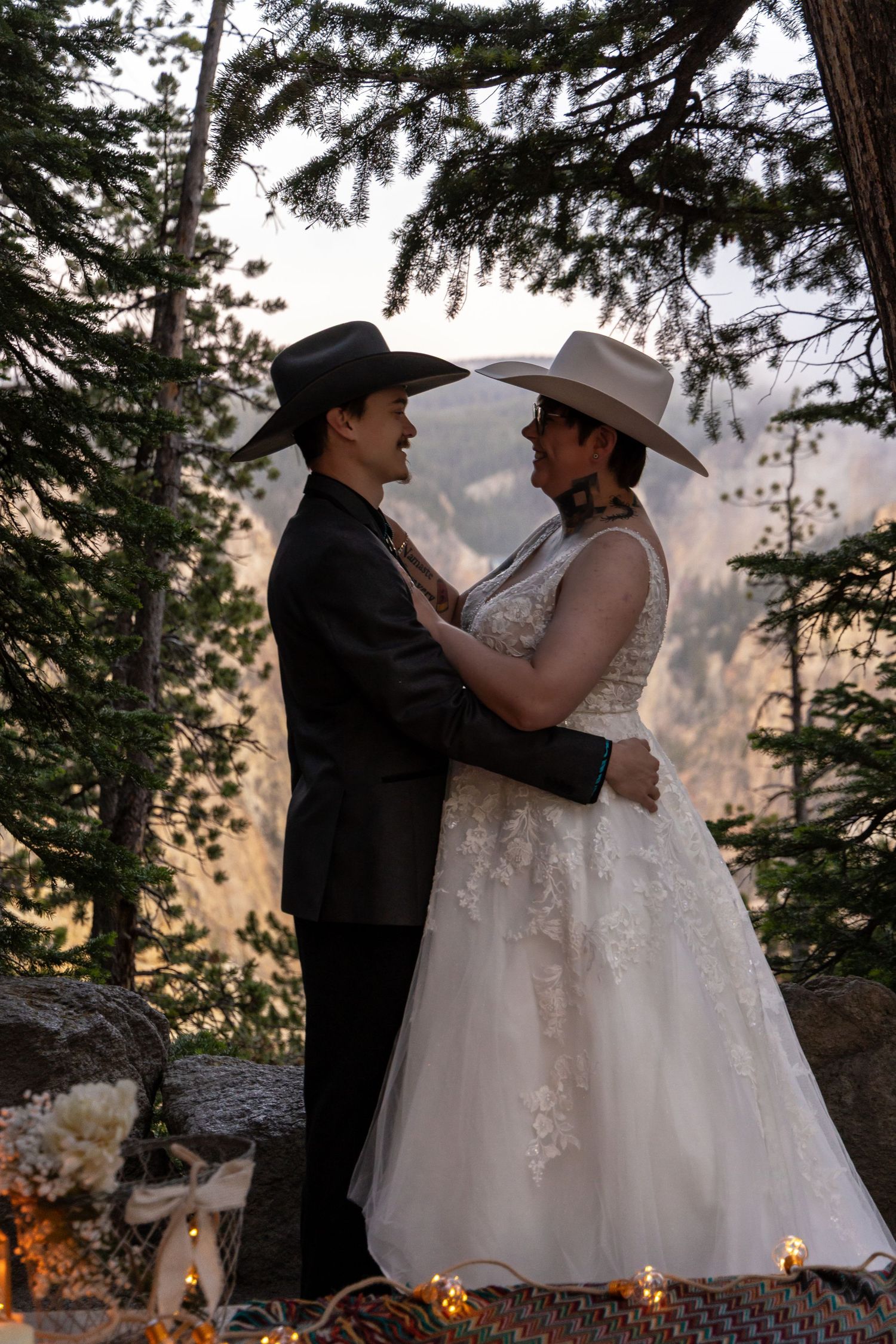 A romantic outdoor wedding scene with a couple in western attire and cowboy hats surrounded by pine trees at sunset.