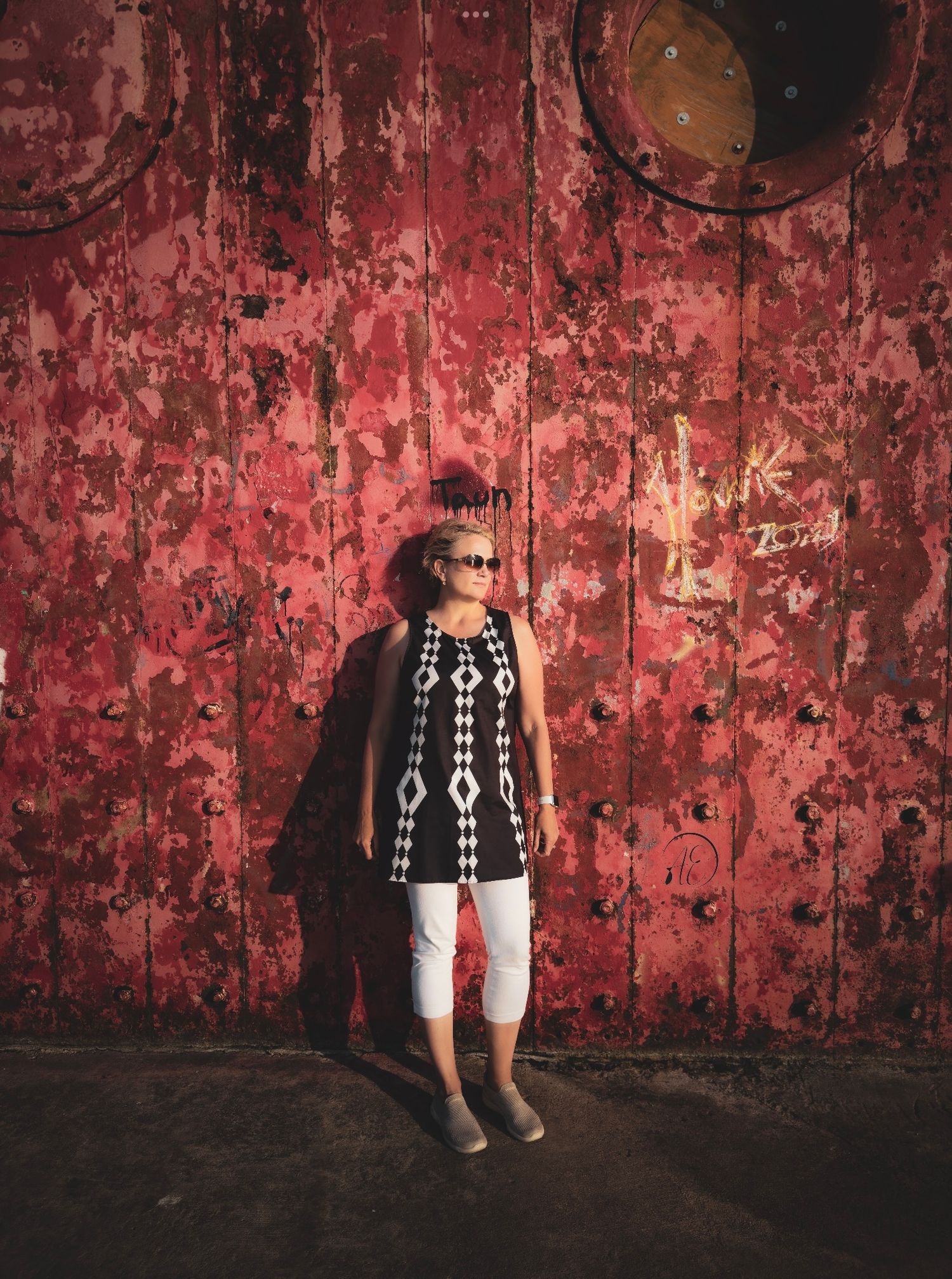 Woman in black and white top and white pants standing against a rustic red peeling paint metal wall.