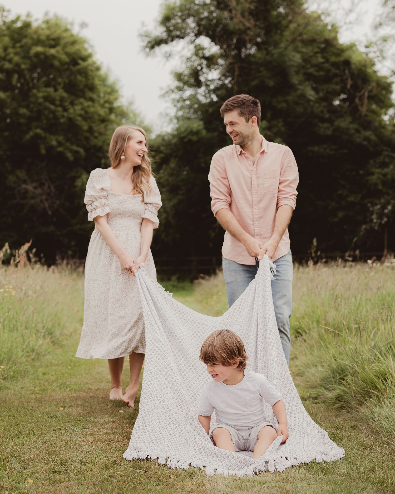 Smiling couple in a meadow lifting a white blanket over their toddler son sitting on the ground.