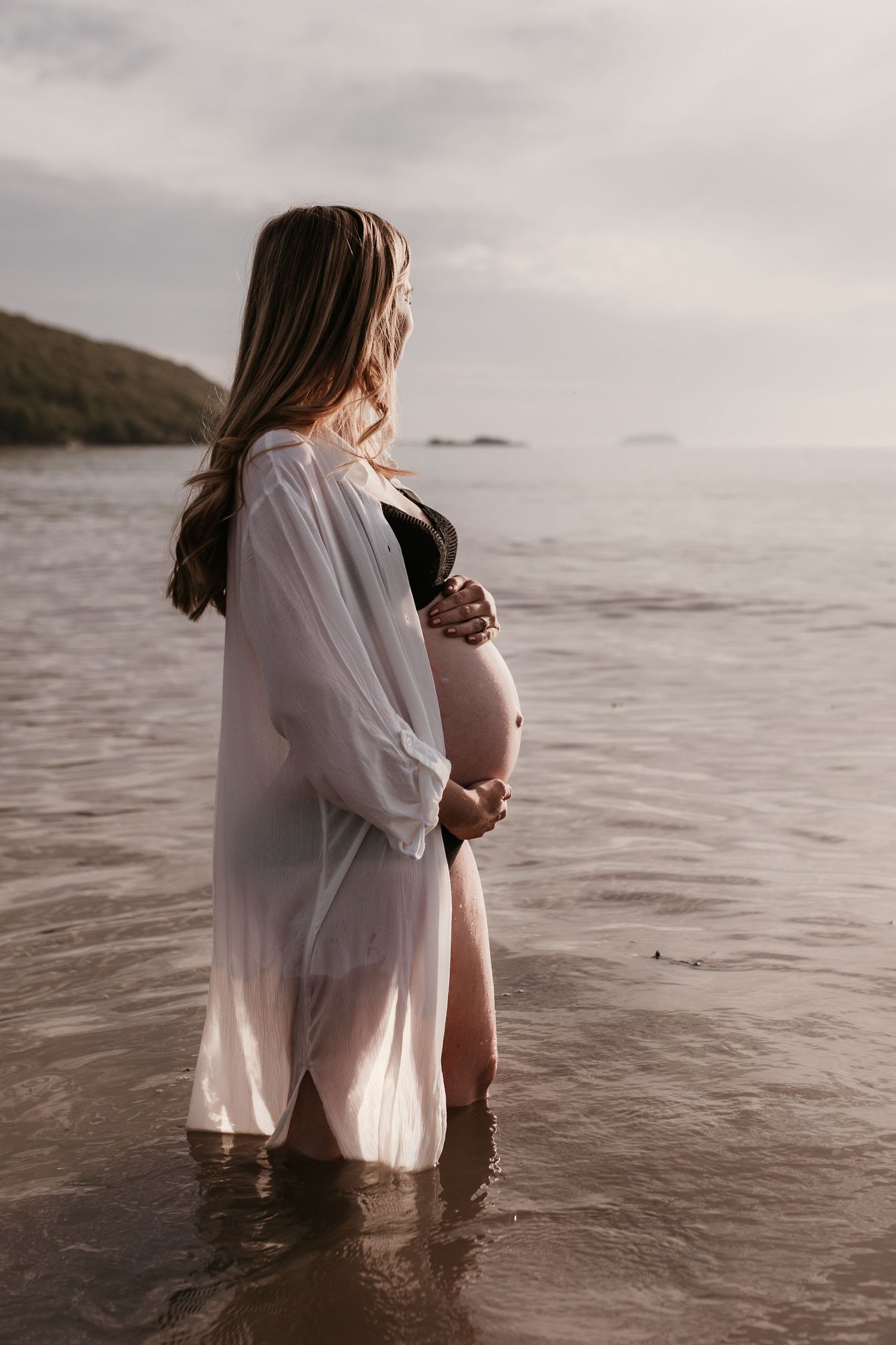 Pregnant woman in white sheer robe standing in shallow ocean water, cradling her baby bump while gazing at the sea.