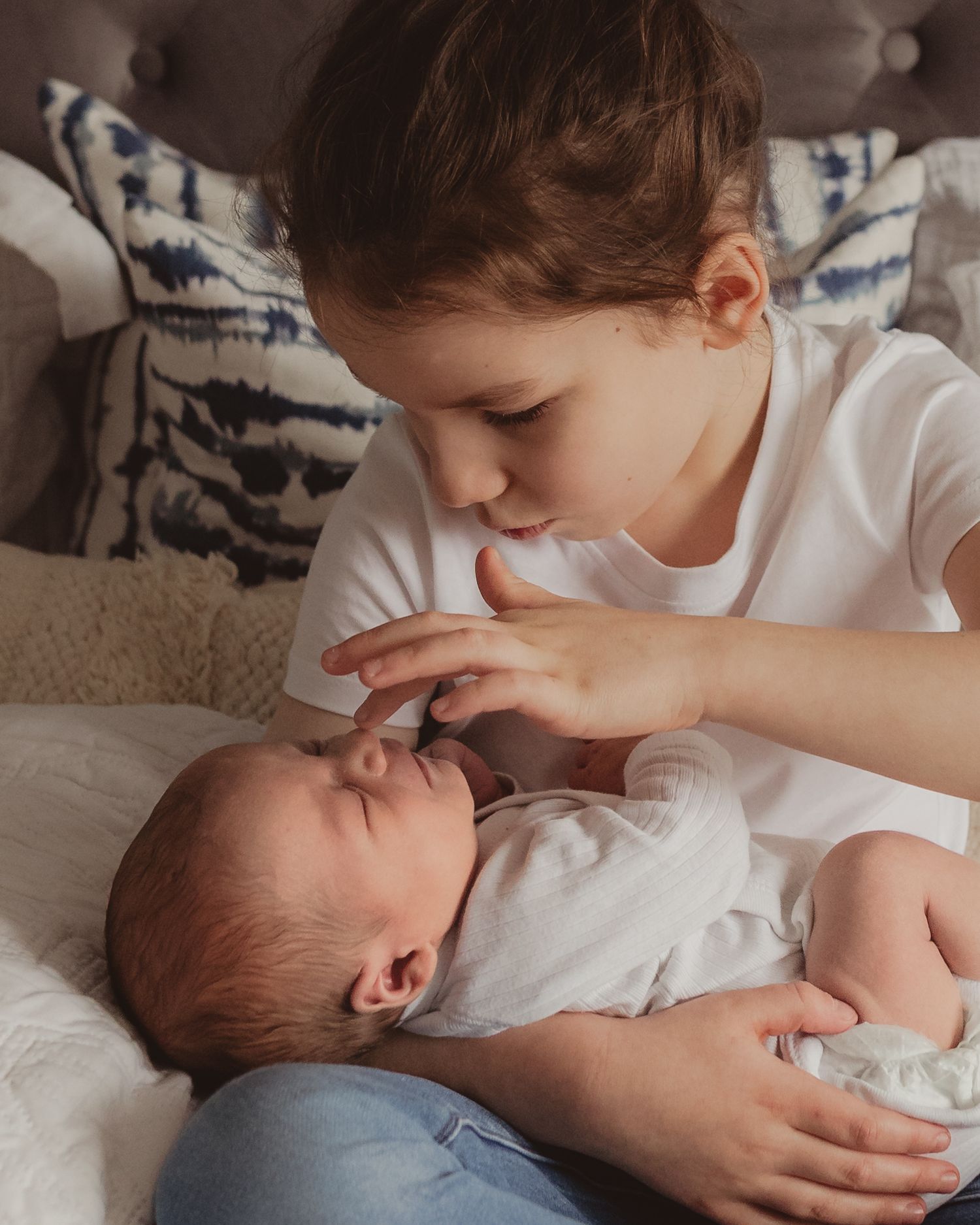 Young child tenderly kissing a sleeping newborn baby while cradling them on a bed.