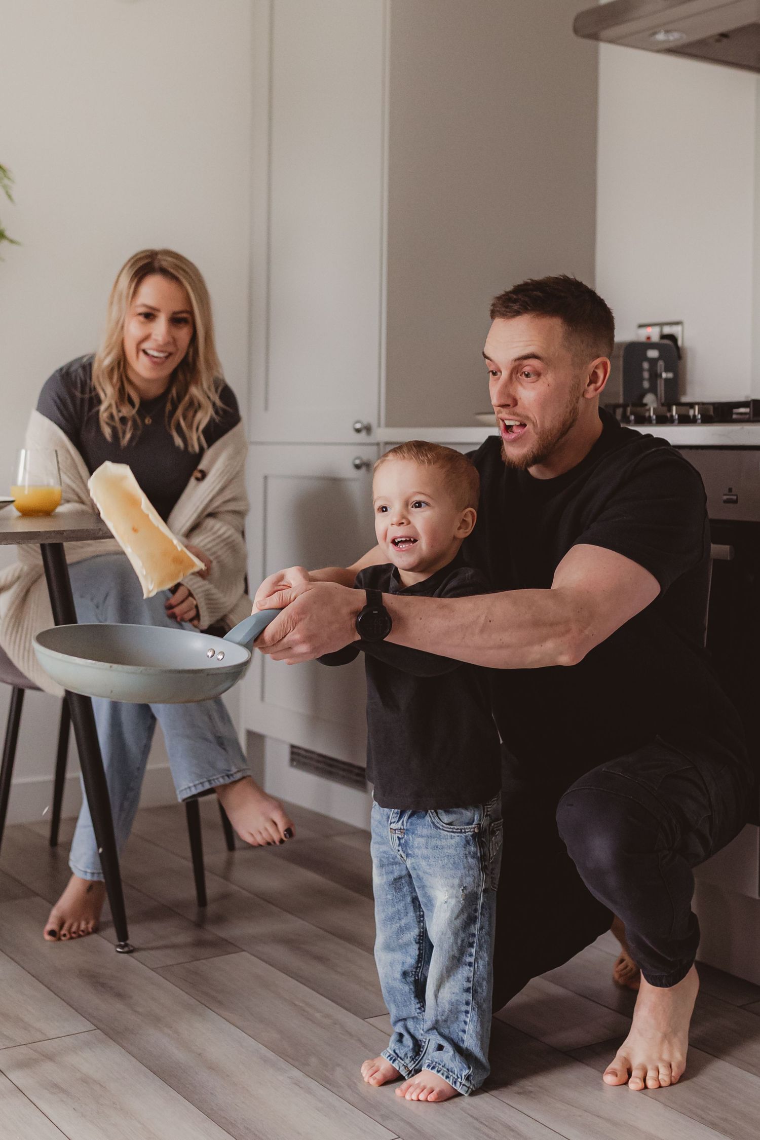 Happy family in kitchen, father teaching toddler son while smiling mother sits nearby holding a juice glass.