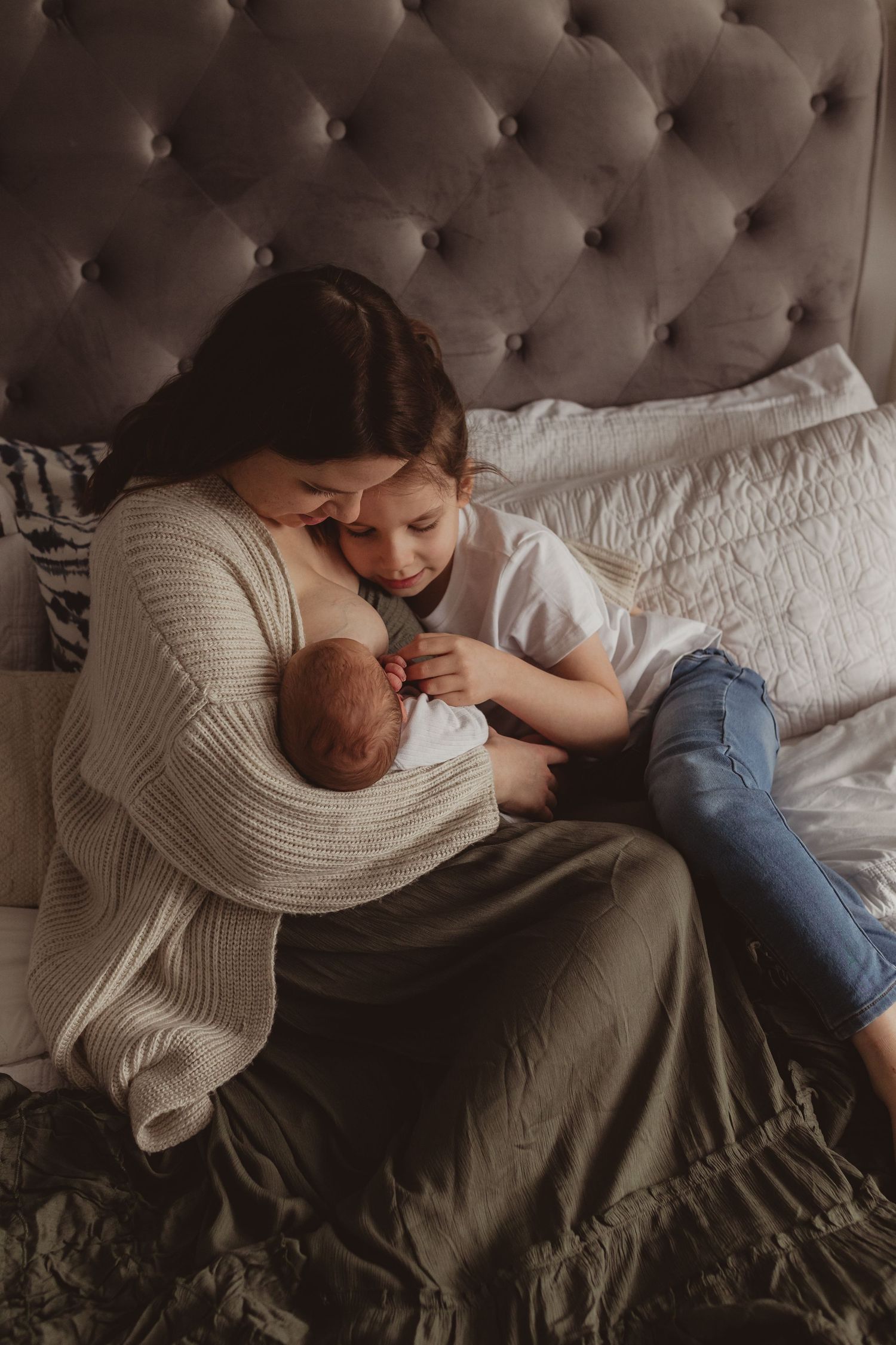 A mother in a cream sweater holds her newborn on a bed while an older child lovingly leans in close.